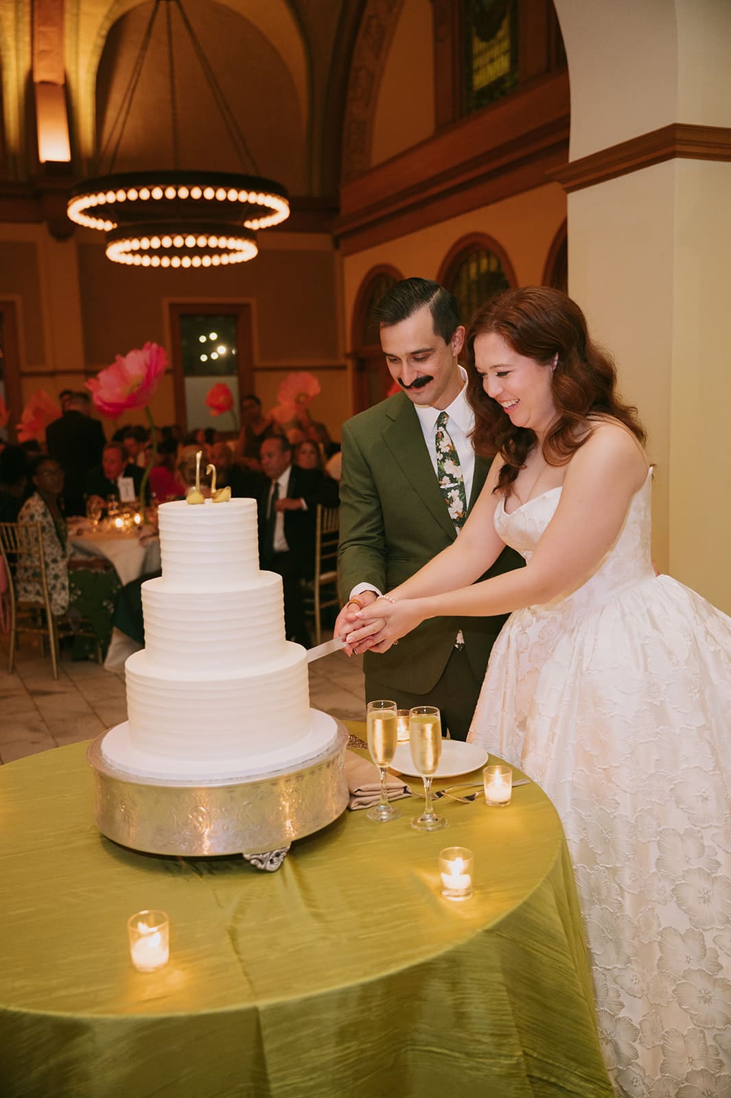A bride and groom cut a four-tier white wedding cake together at a reception, surrounded by guests in a decorated hall.