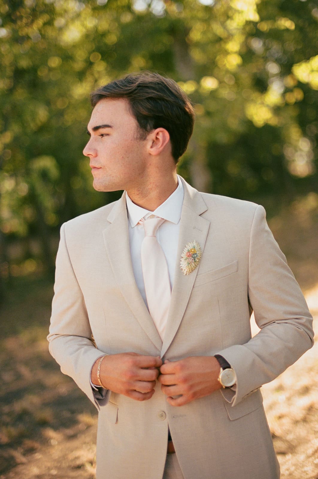 A man in a light beige suit and white shirt stands outdoors, looking to the side with trees blurred in the background.