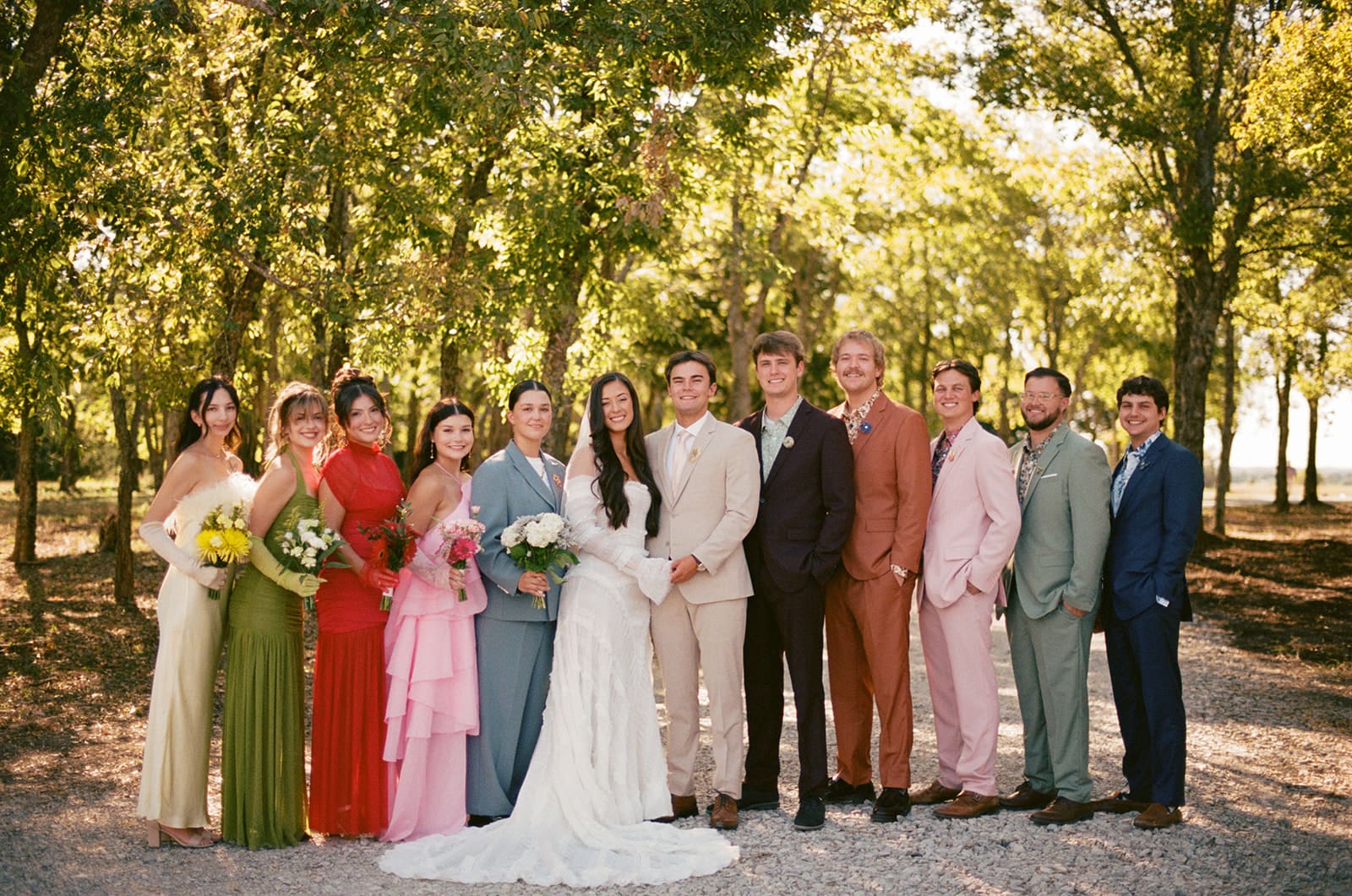 A bride and groom stand together outdoors, surrounded by their wedding party dressed in colorful formal attire, smiling and laughing.