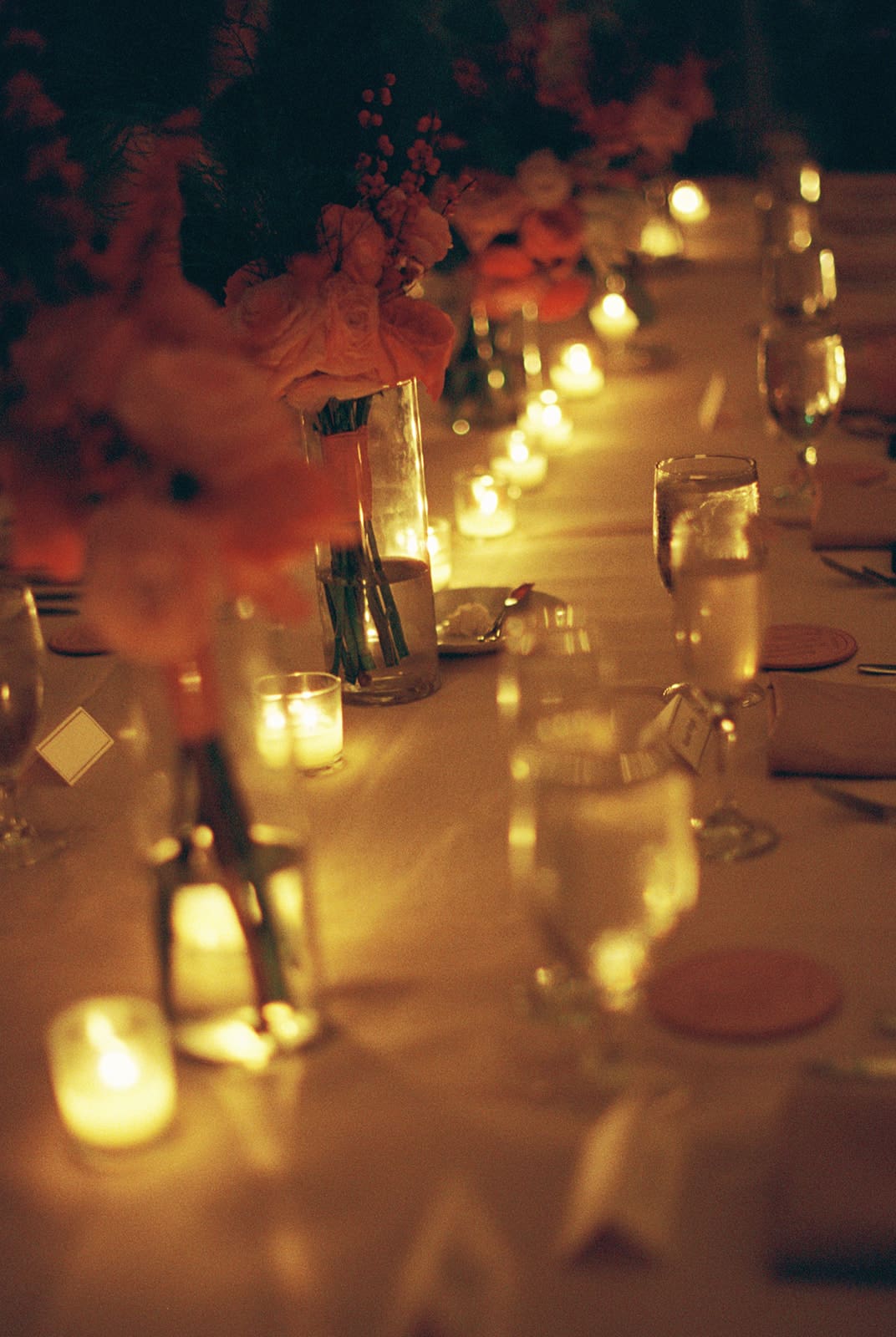 A banquet table set with glassware, plates, flower arrangements, and lit candles creating a warm, ambient glow.