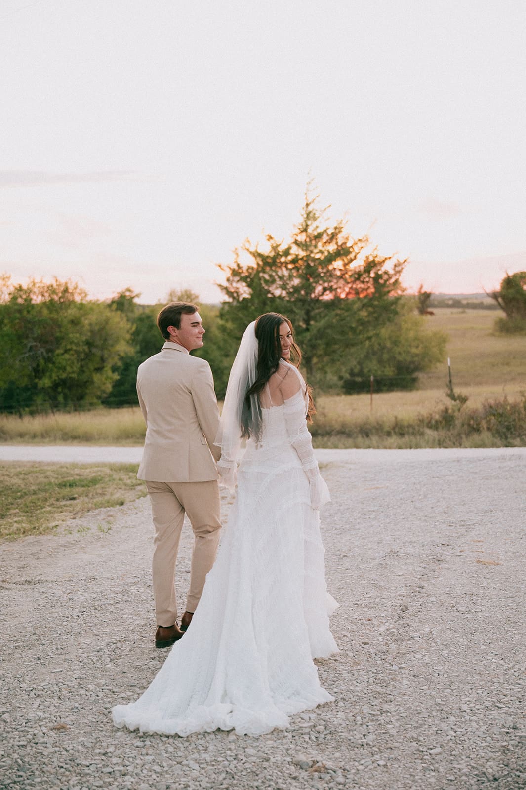A bride in a white dress and veil stands beside a groom in a beige suit outdoors on a gravel path, with trees and fields in the background for a backyard wedding in texas