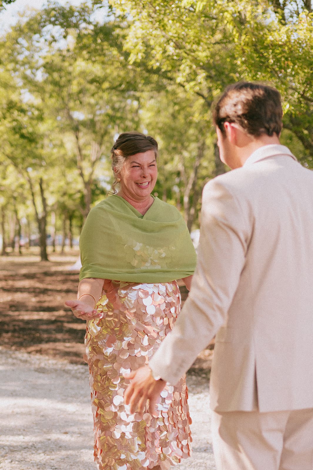 A woman in a green shawl and sequin skirt smiles at a man in a light suit outdoors on a sunny day.
