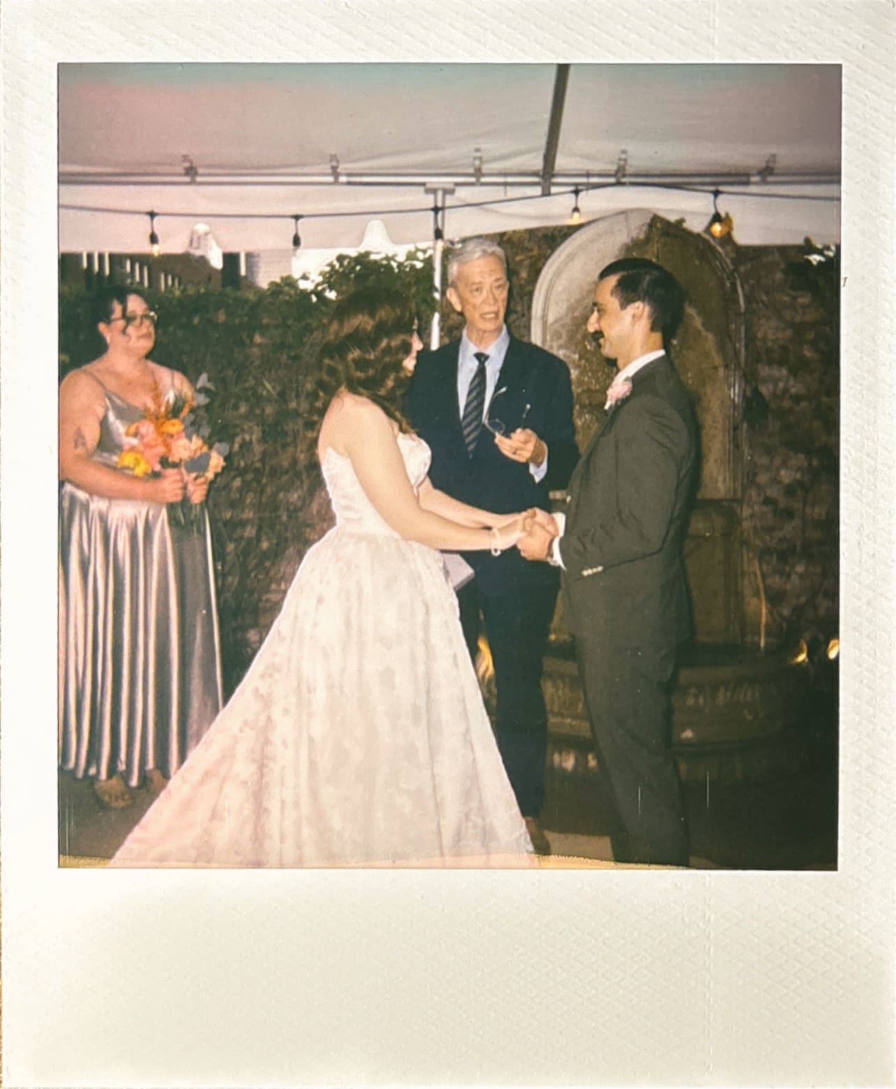A bride and groom stand holding hands during their wedding ceremony, with an officiant and bridesmaids present, under a tent with string lights at The Ashton Depot in Fort Worth