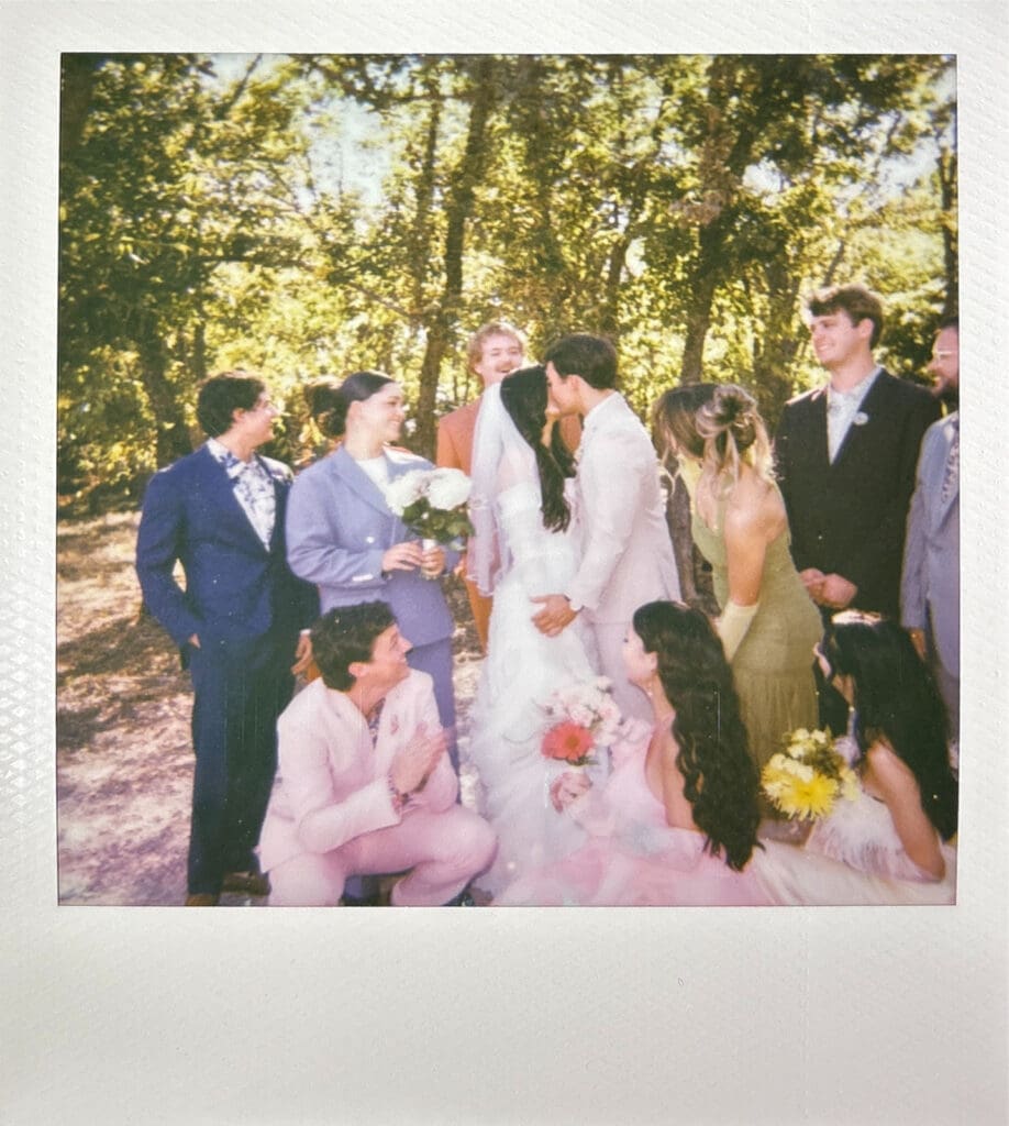 A bride and groom kiss outdoors, surrounded by their wedding party dressed in formal attire, with trees in the background.