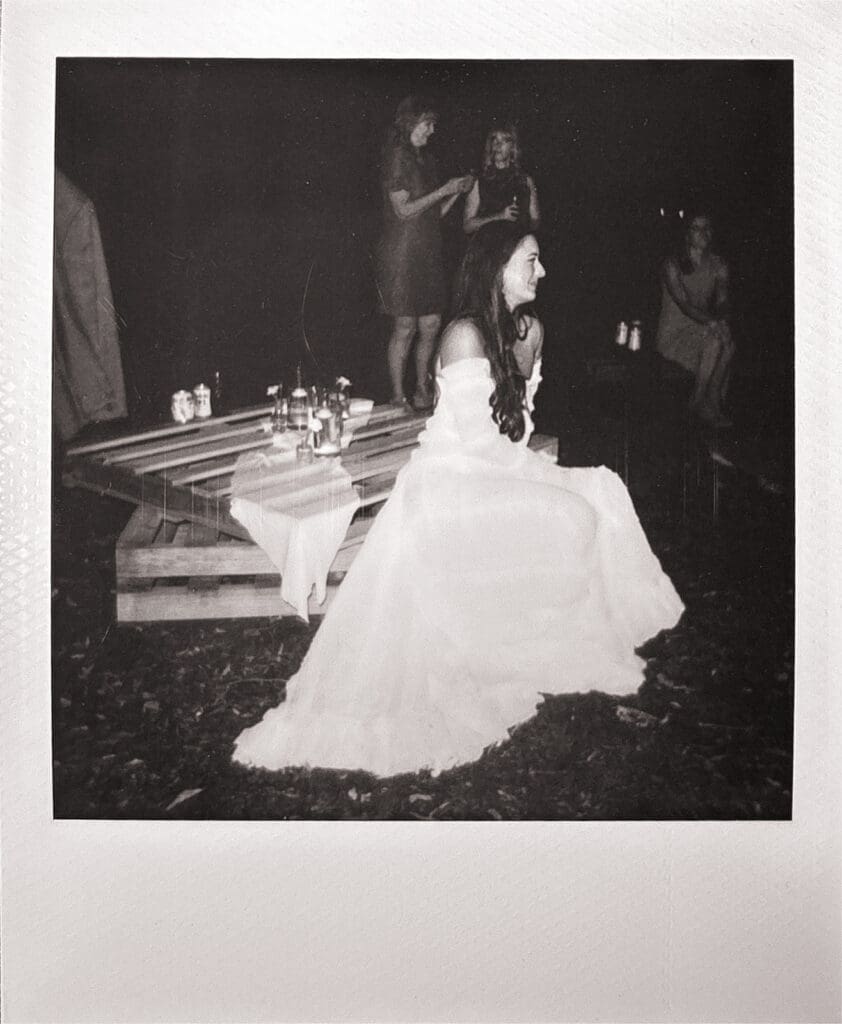 A woman in a white dress sits on a blanket-covered bench outdoors at night, with three people standing and sitting in the background for a backyard wedding in texas