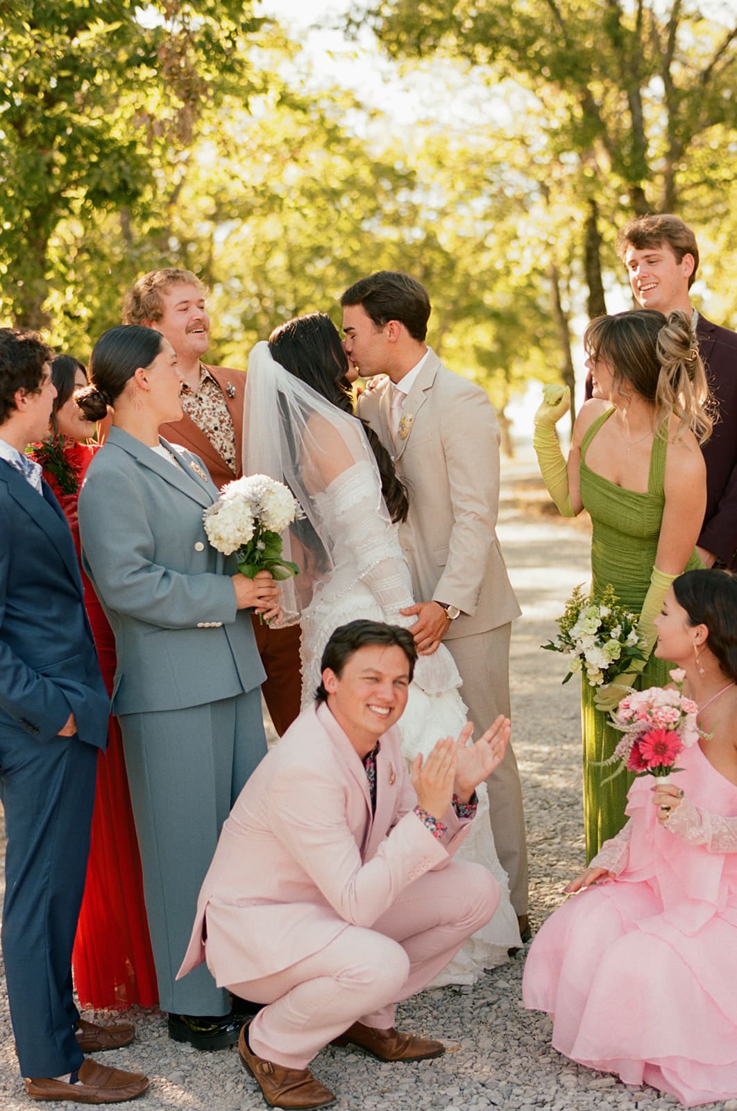 A bride and groom stand together outdoors, surrounded by their wedding party dressed in colorful formal attire, smiling and laughing.