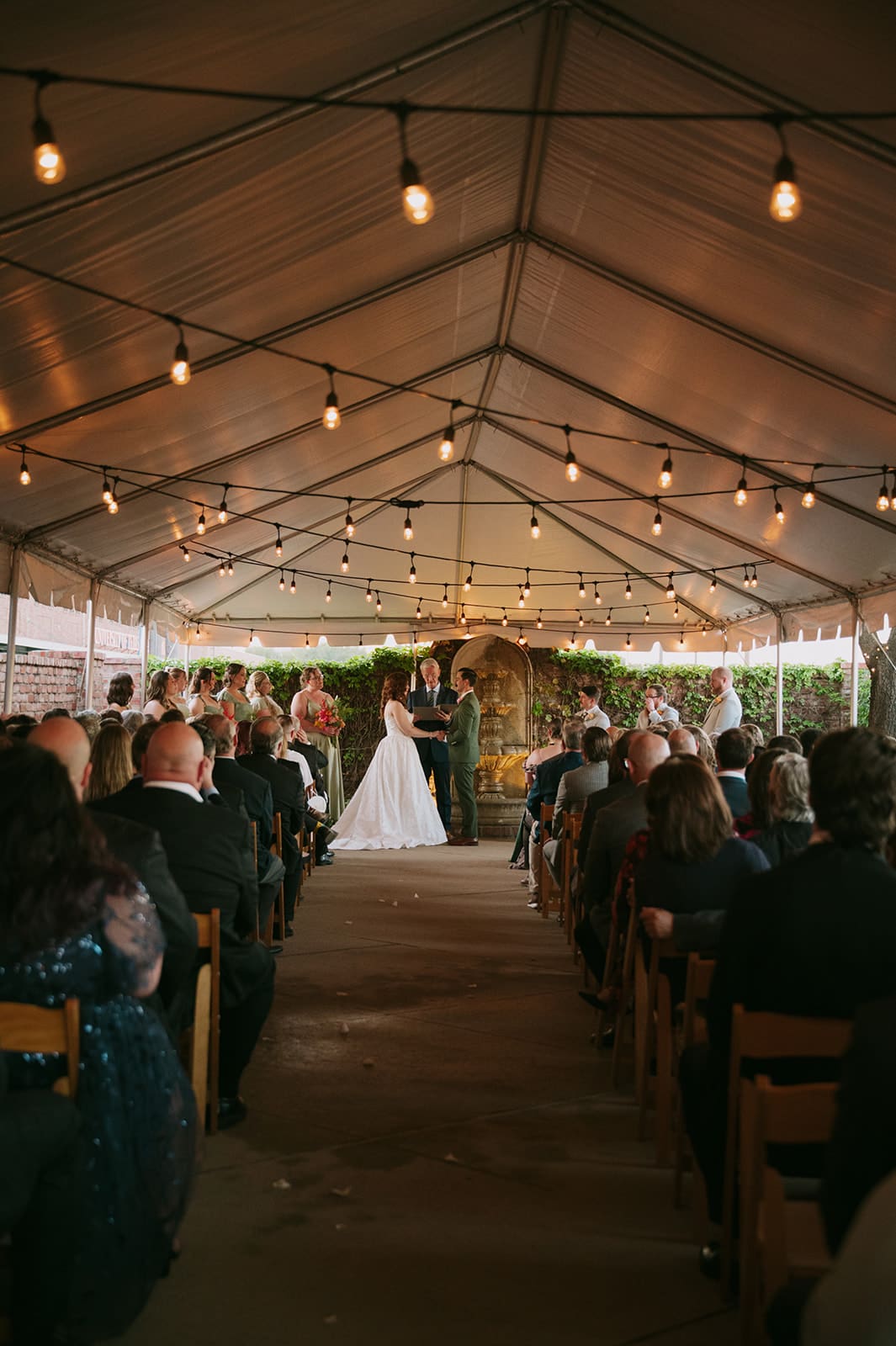 A bride and groom stand holding hands during their wedding ceremony, with an officiant and bridesmaids present, under a tent with string lights at The Ashton Depot in Fort Worth
