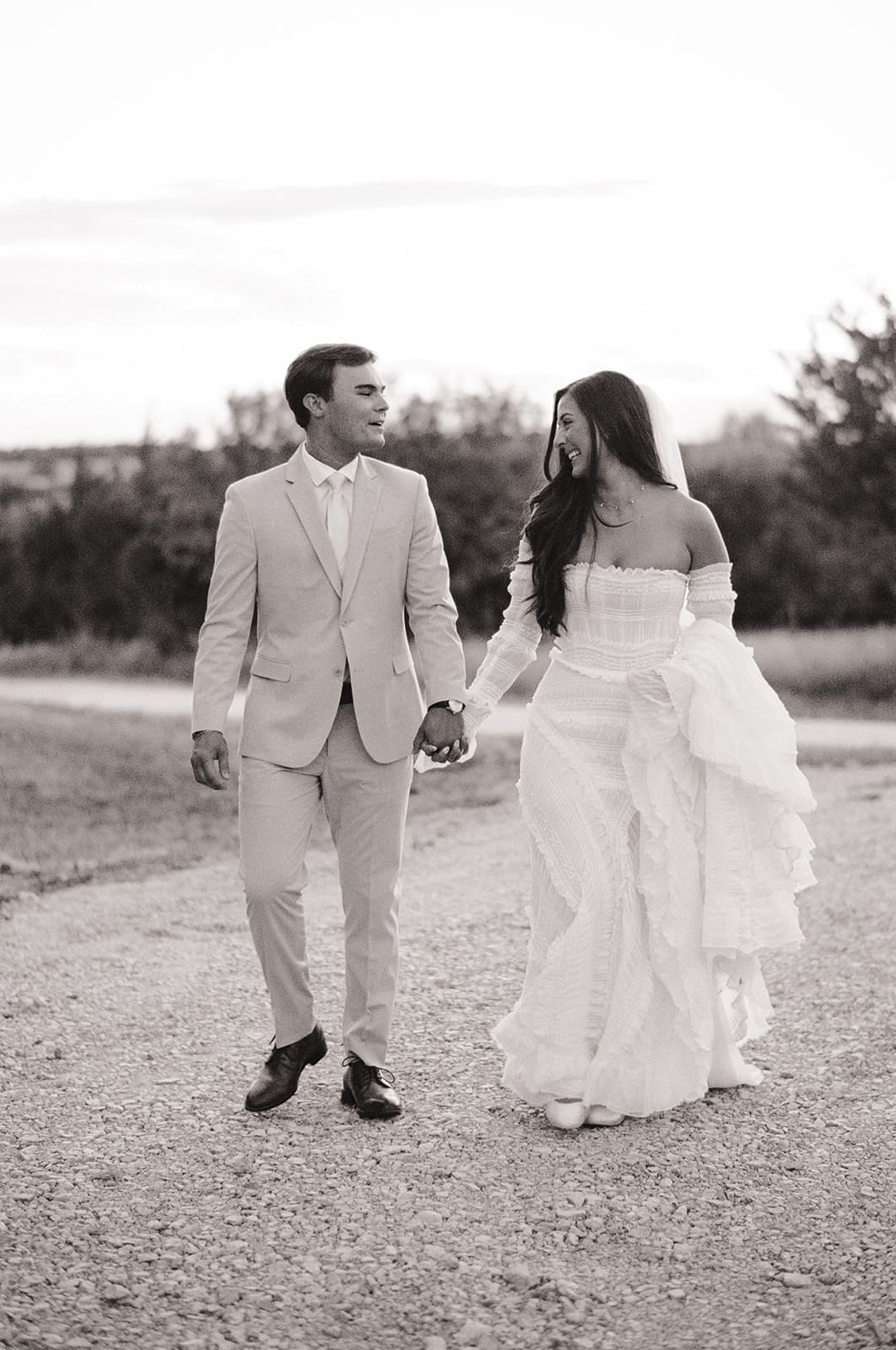 A bride and groom kiss outdoors at sunset; the bride shows her ring while wearing a white dress and the groom wears a beige suite at their backyard wedding