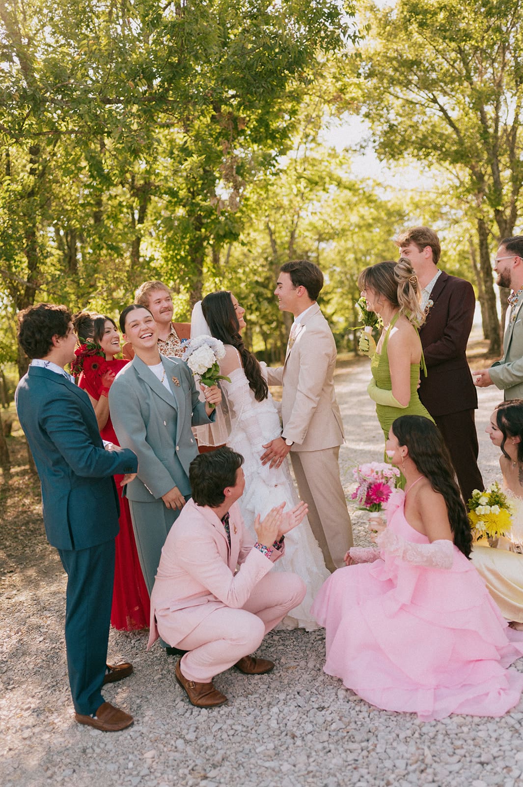 A bride and groom stand together outdoors, surrounded by their wedding party dressed in colorful formal attire, smiling and laughing.
