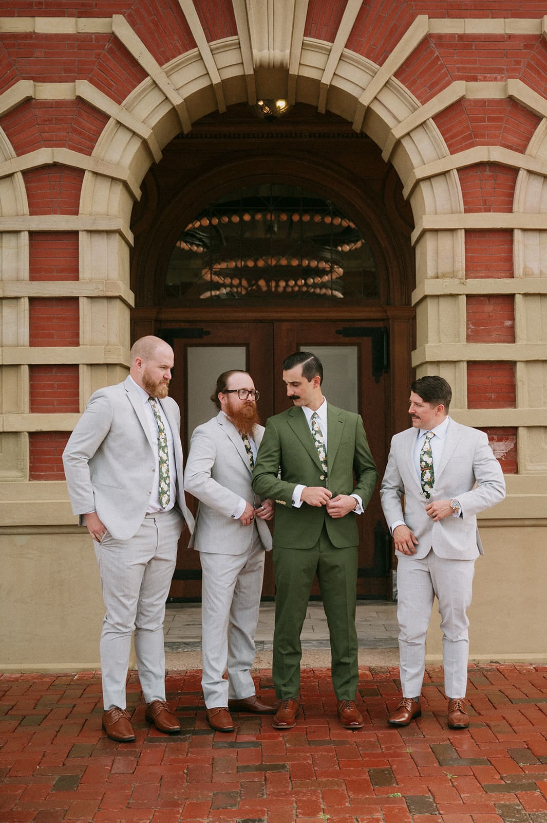 Four men in suits stand in front of an arched brick building entrance; three wear light gray suits and one wears a green suit.
