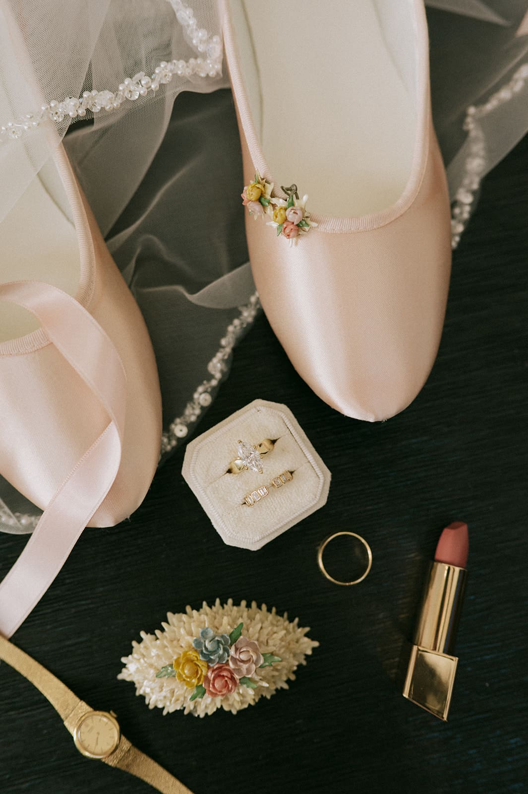 Flat lay of wedding items including a wedding invitation, a pink shoe, three rings, a gold watch, lipstick, veil, and a decorated envelope.