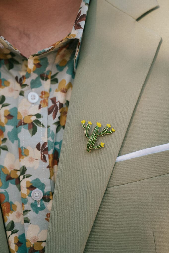A man in a light beige suit and white shirt stands outdoors, looking to the side with trees blurred in the background.
