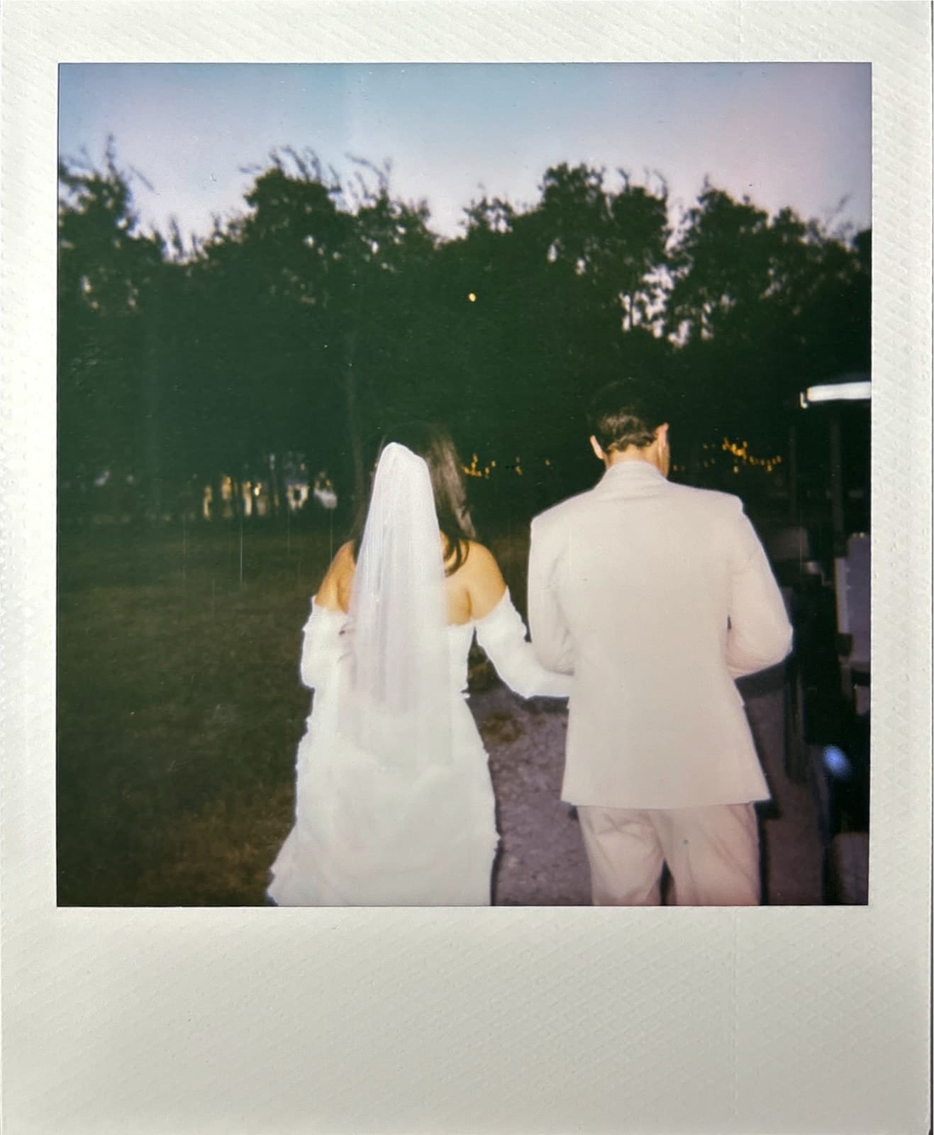 A bride in a white dress and veil walks arm in arm with a man in a light-colored suit outdoors, facing away from the camera. Trees are visible in the background for a backyard wedding in Texas