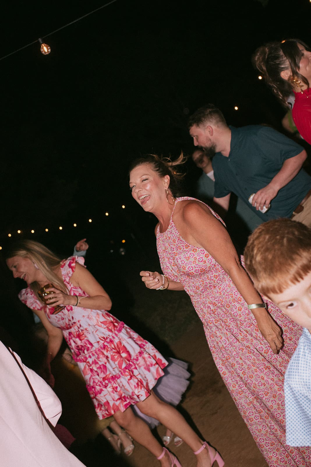 A group of people, including women in floral dresses, smile and dance outdoors at night under string lights