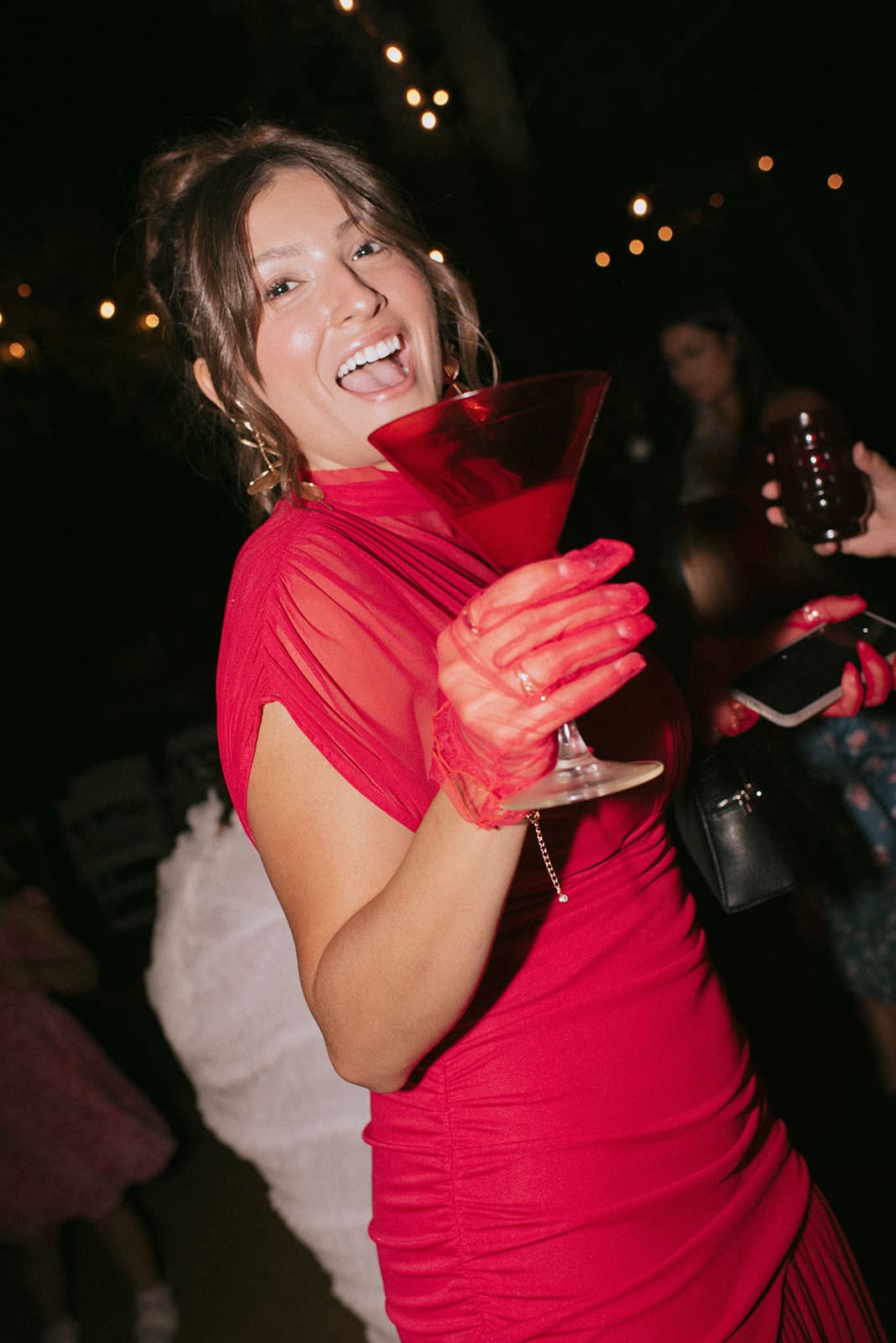 A group of people, including women in floral dresses, smile and dance outdoors at night under string lights for a backyard wedding 