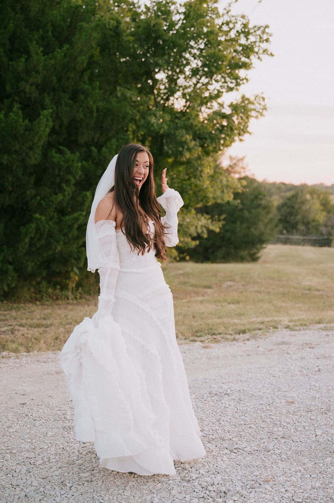 A bride and groom kiss outdoors at sunset; the bride shows her ring while wearing a white dress and the groom wears a beige suite at their backyard wedding