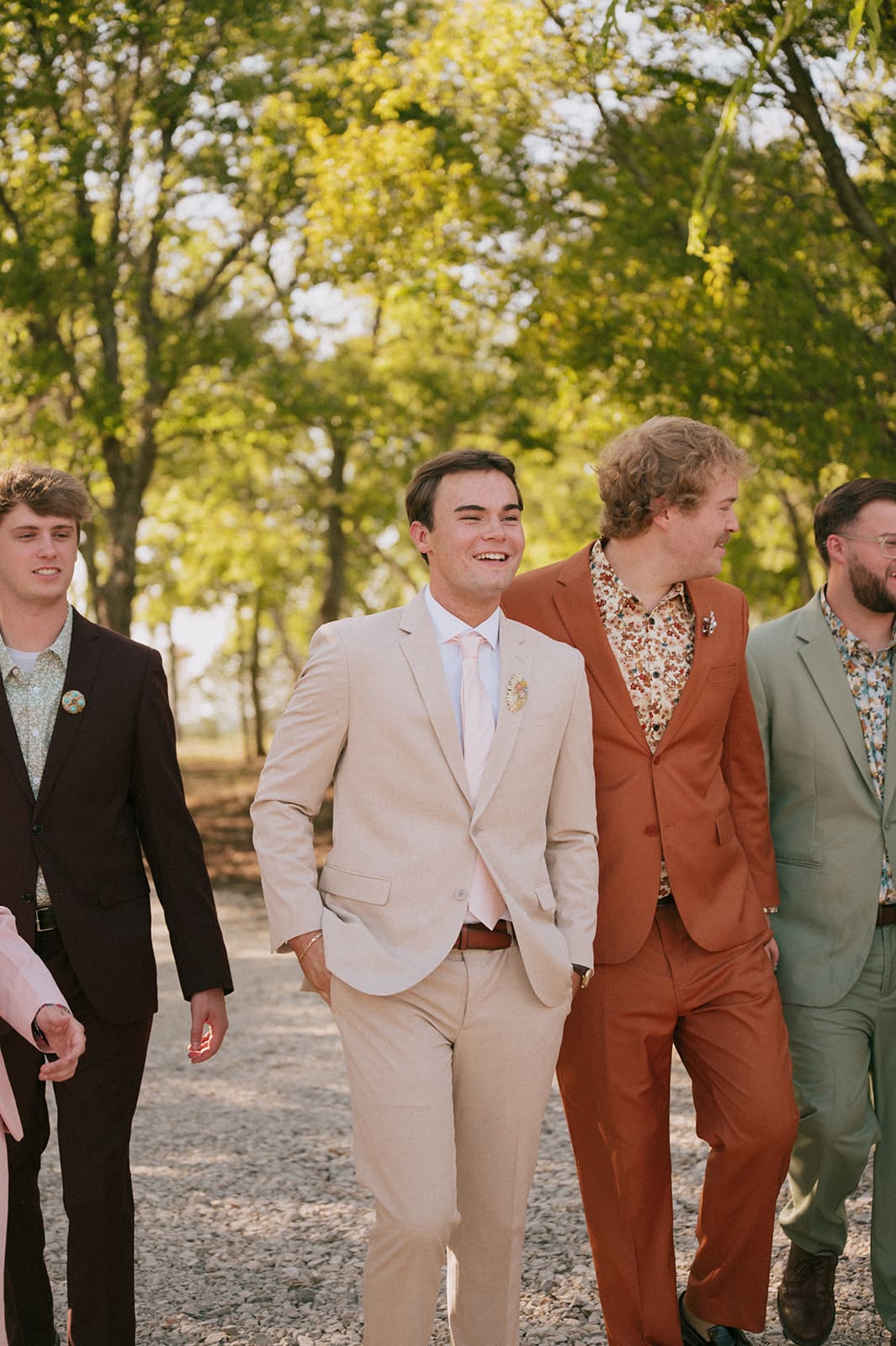 Five men in suits walk outdoors on a gravel path with trees in the background, smiling and interacting with each other.