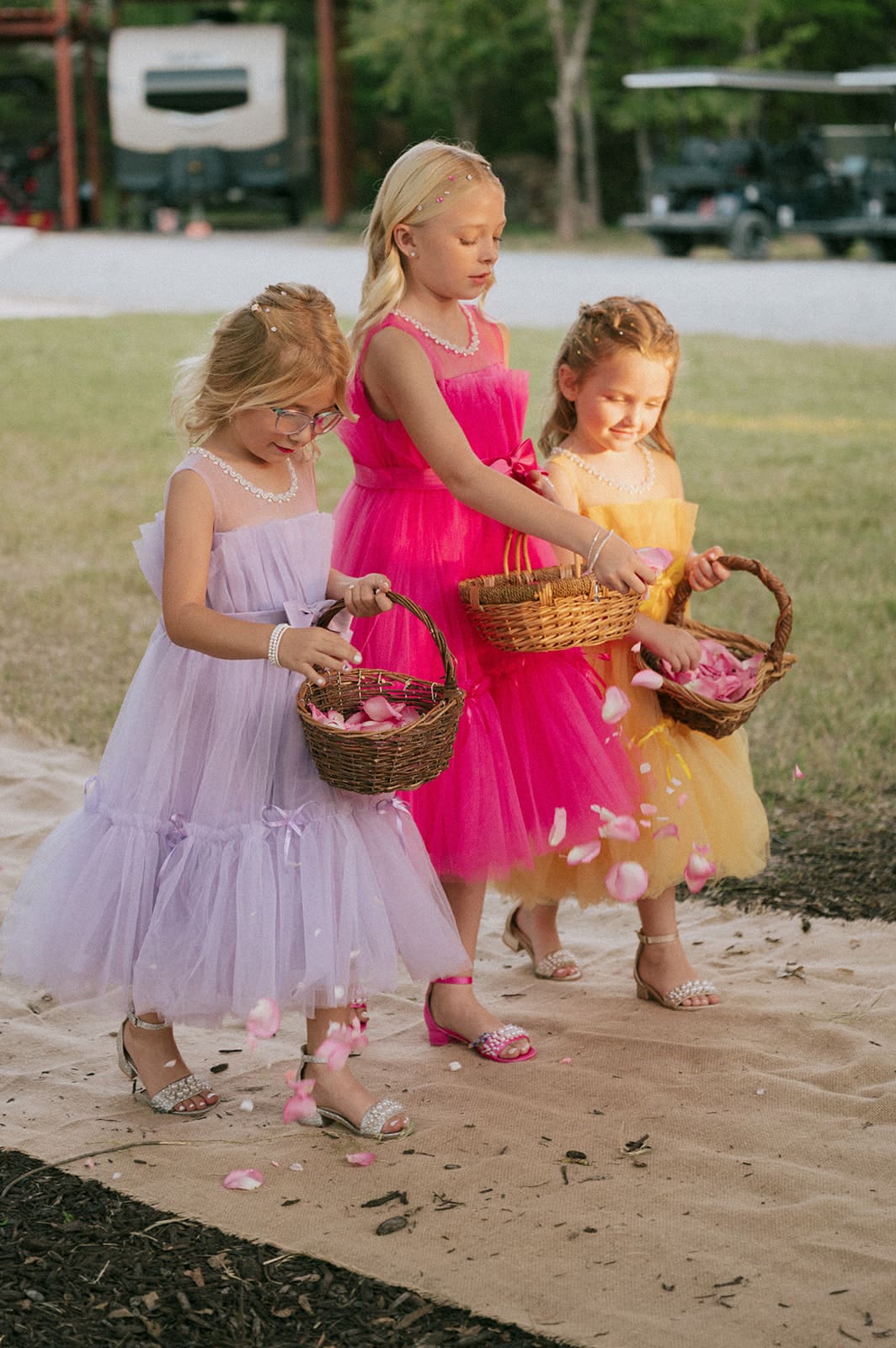 Three young girls in pastel dresses walk on a path, scattering flower petals from baskets during an outdoor event.