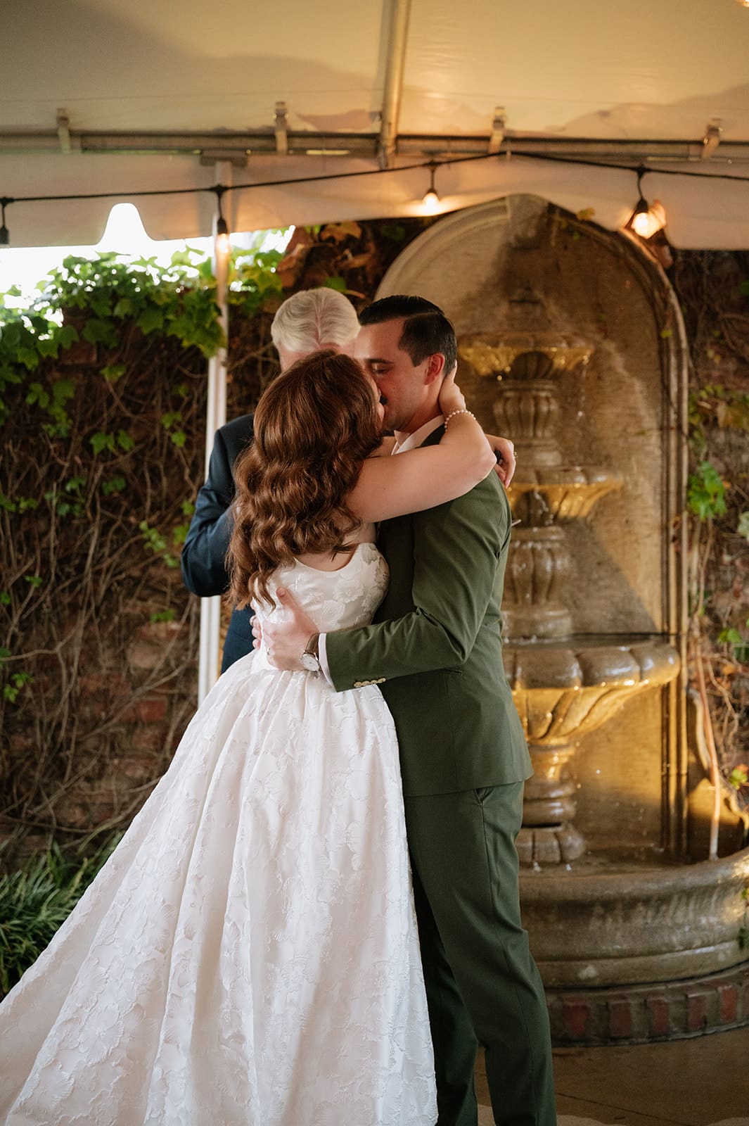 A bride and groom stand holding hands during their wedding ceremony, with an officiant and bridesmaids present, under a tent with string lights at The Ashton Depot in Fort Worth