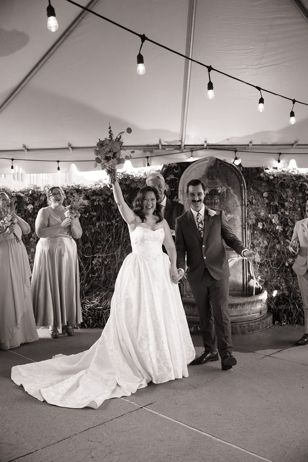 A bride and groom stand holding hands during their wedding ceremony, with an officiant and bridesmaids present, under a tent with string lights at The Ashton Depot in Fort Worth