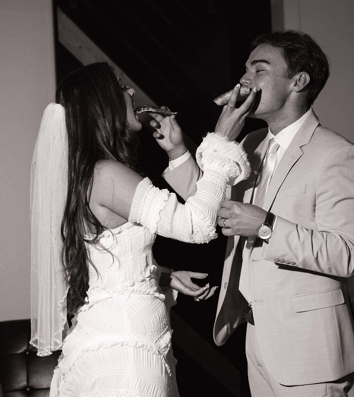 A bride and groom in wedding attire feed each other cake, smiling and standing close together.