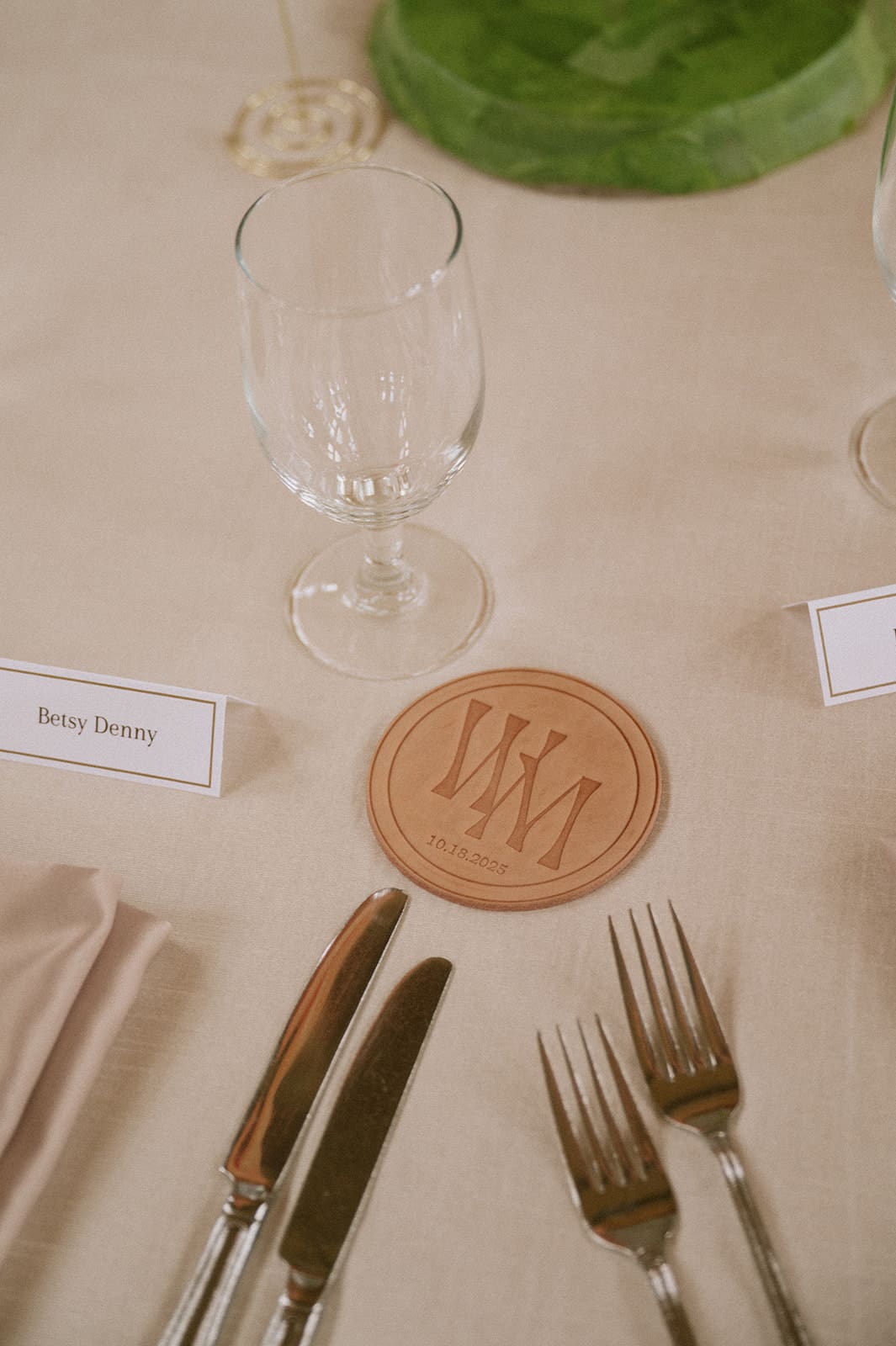 A place setting with two forks, a knife, a spoon, a glass, a napkin, a name card, and a round leather coaster with a monogram on a beige tablecloth.