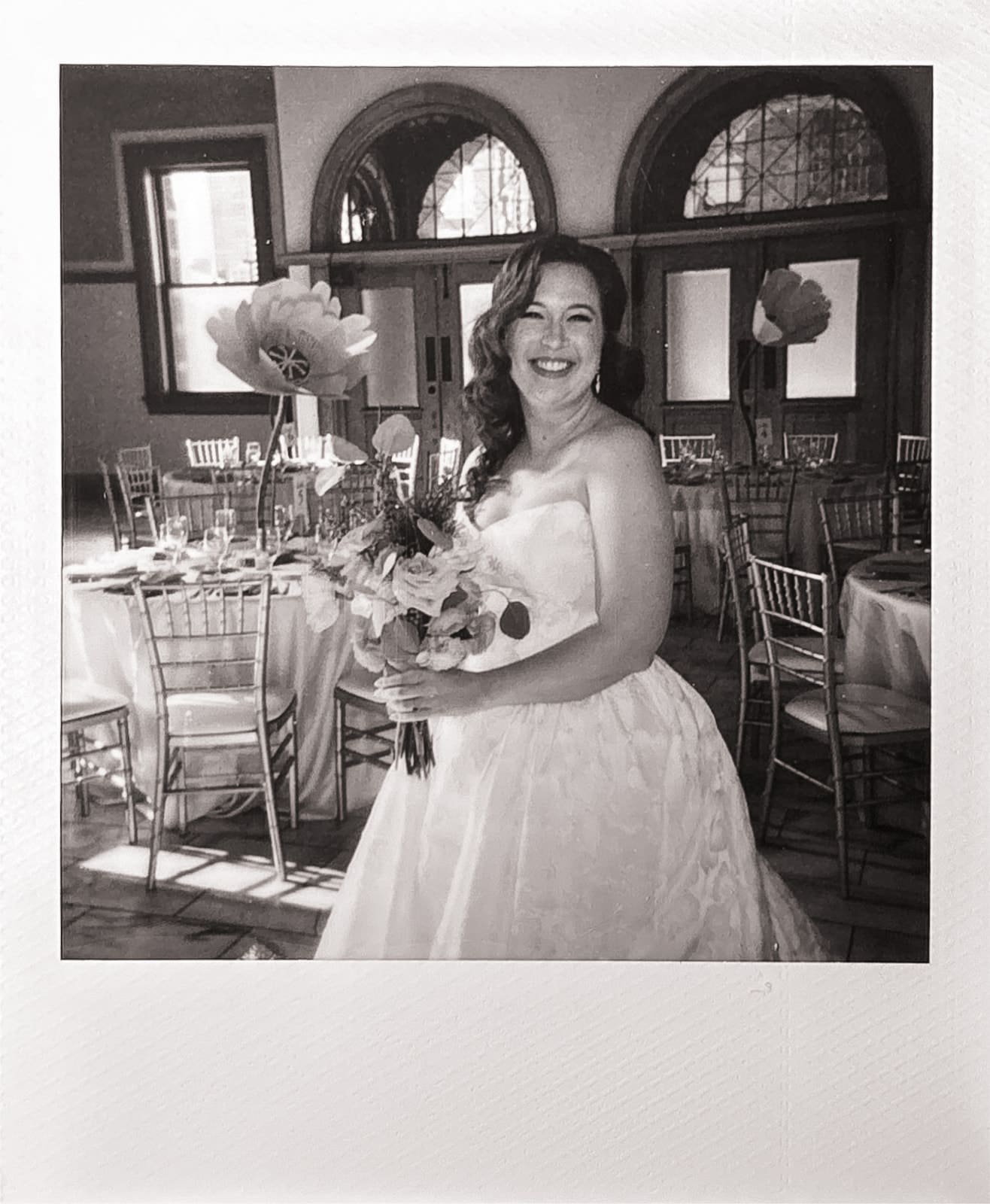 A woman in a strapless white wedding dress holds a bouquet and smiles, standing in a decorated banquet hall with set tables and chairs.