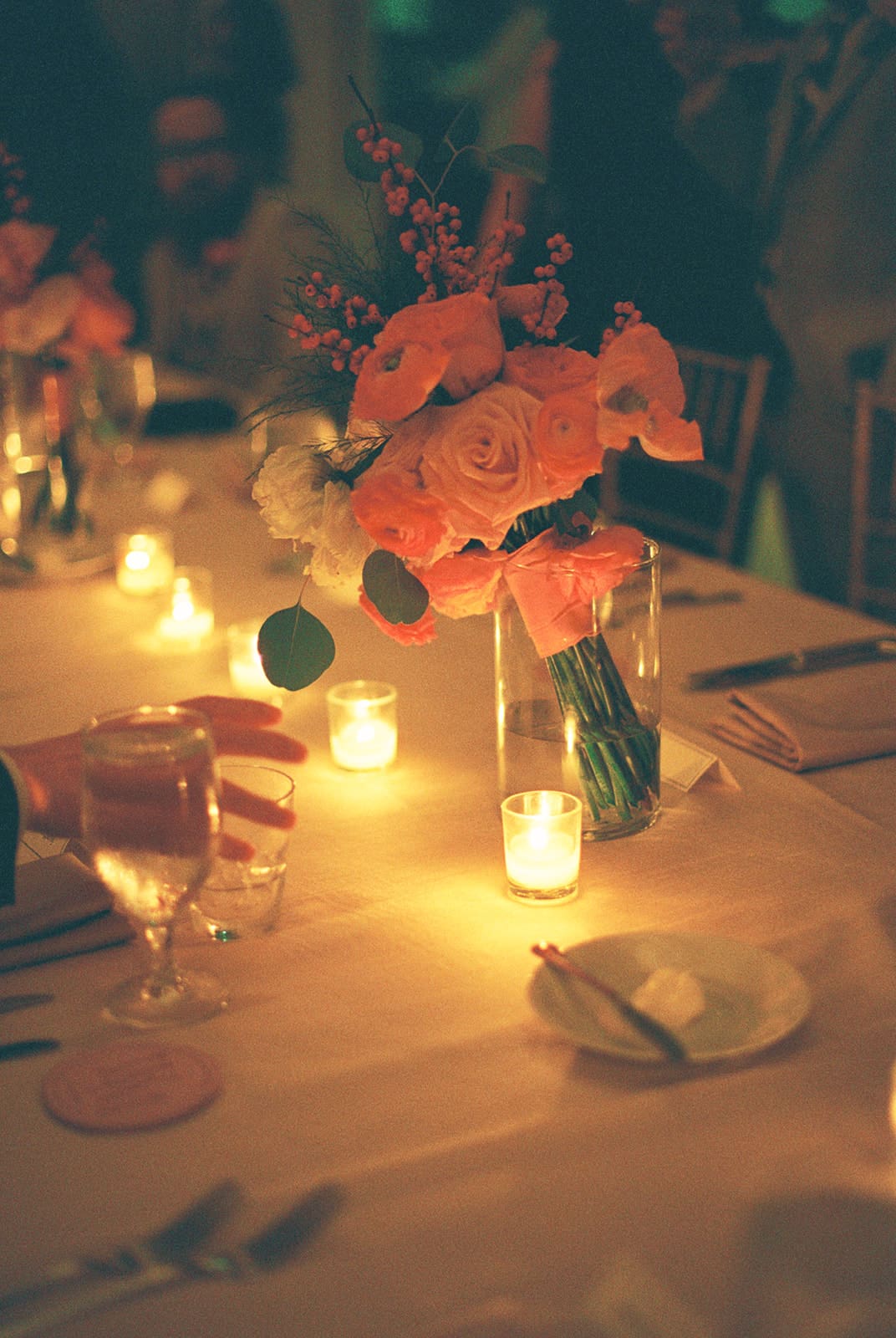 A banquet table set with glassware, plates, flower arrangements, and lit candles creating a warm, ambient glow.