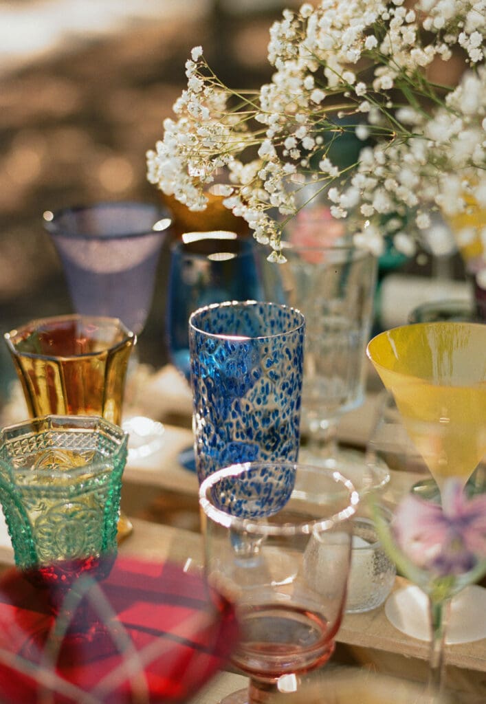 A variety of colorful glass goblets are arranged on a wooden pallet table, decorated with small white flowers, at an outdoor event.
