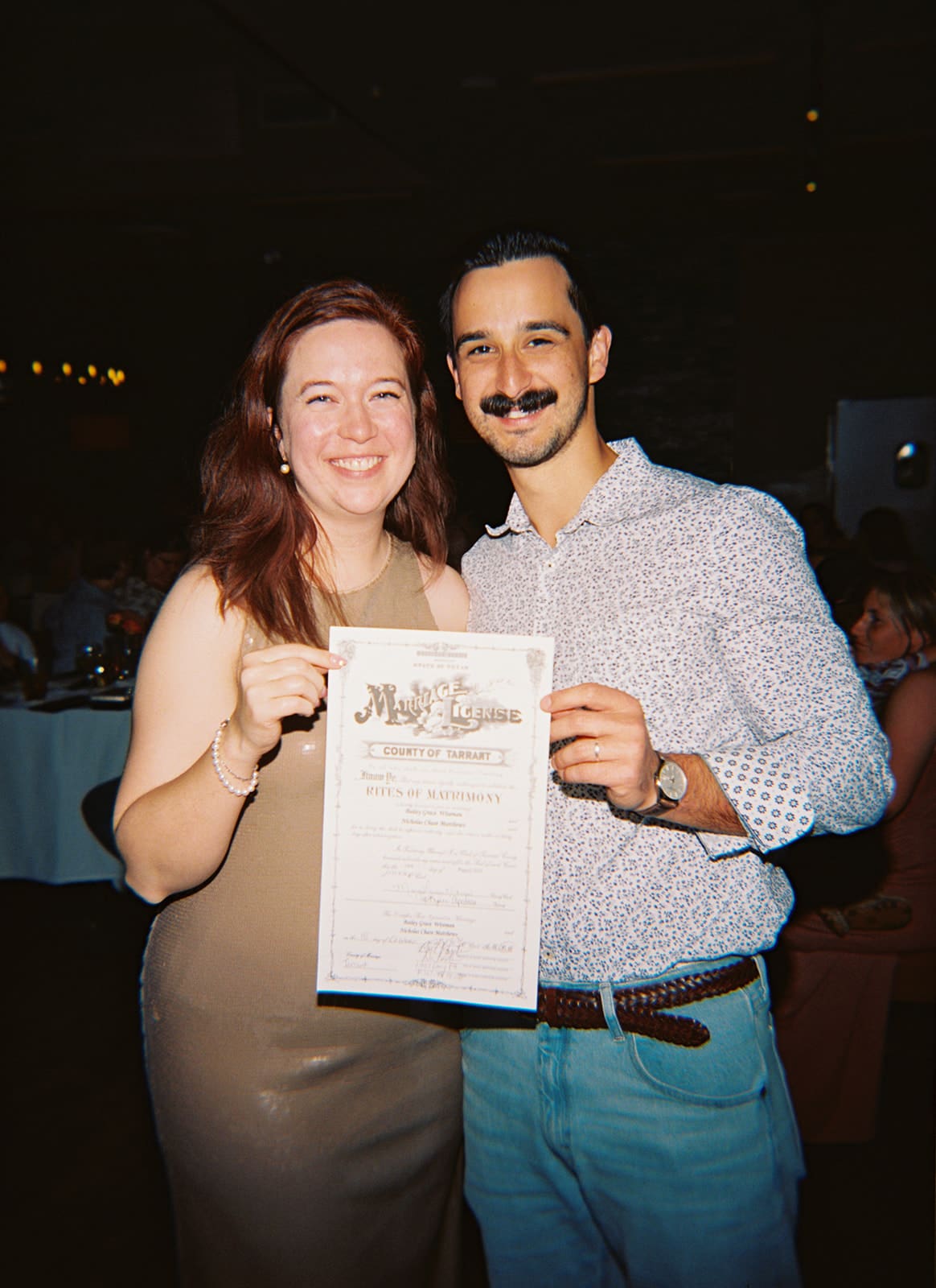 A woman and a man stand together indoors, smiling and holding up a certificate or document toward the camera.