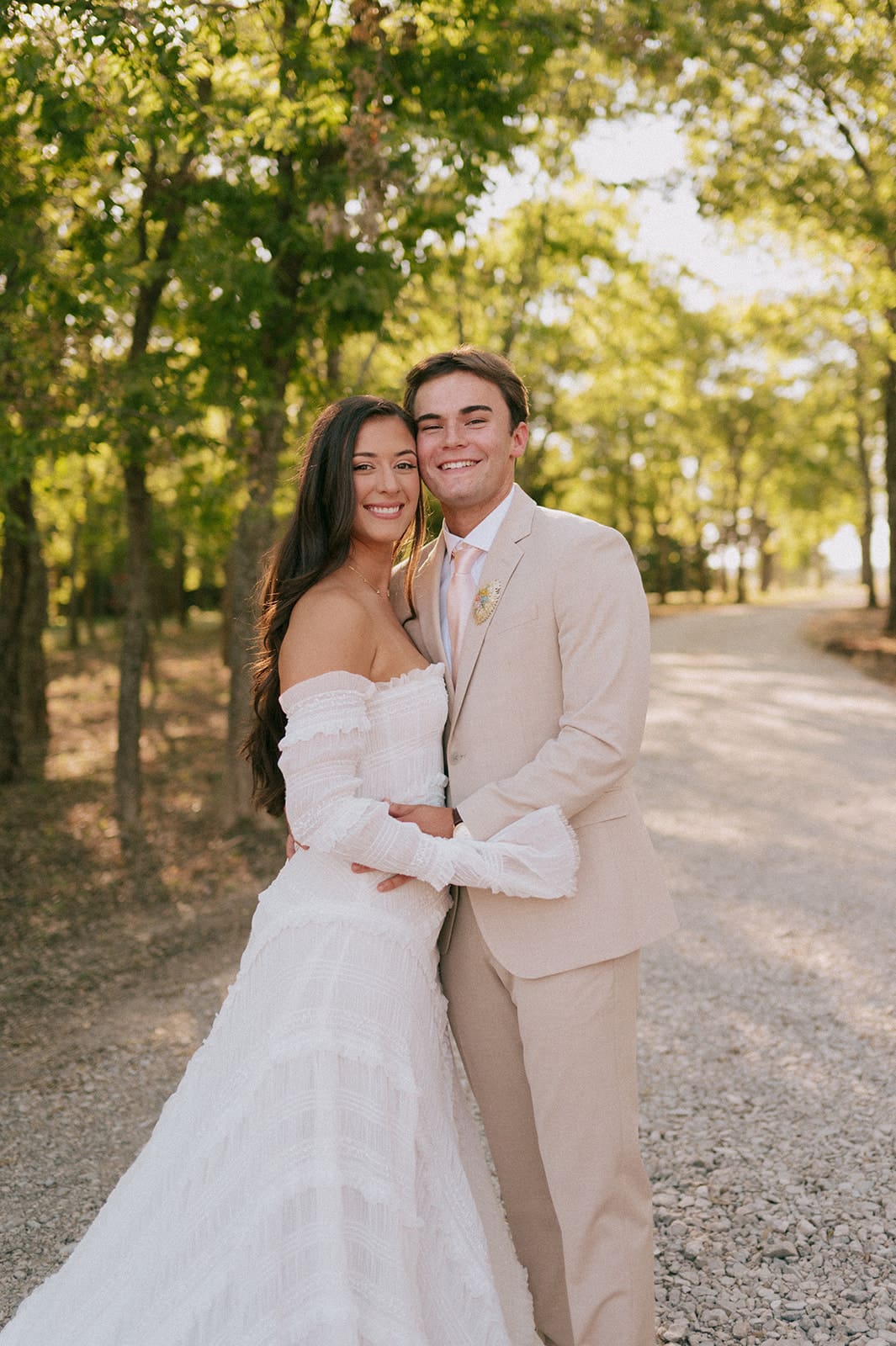 A bride and groom stand outdoors facing each other, holding hands in sunlight, with the bride holding a large bouquet of pink flowers for a backyard wedding in texas