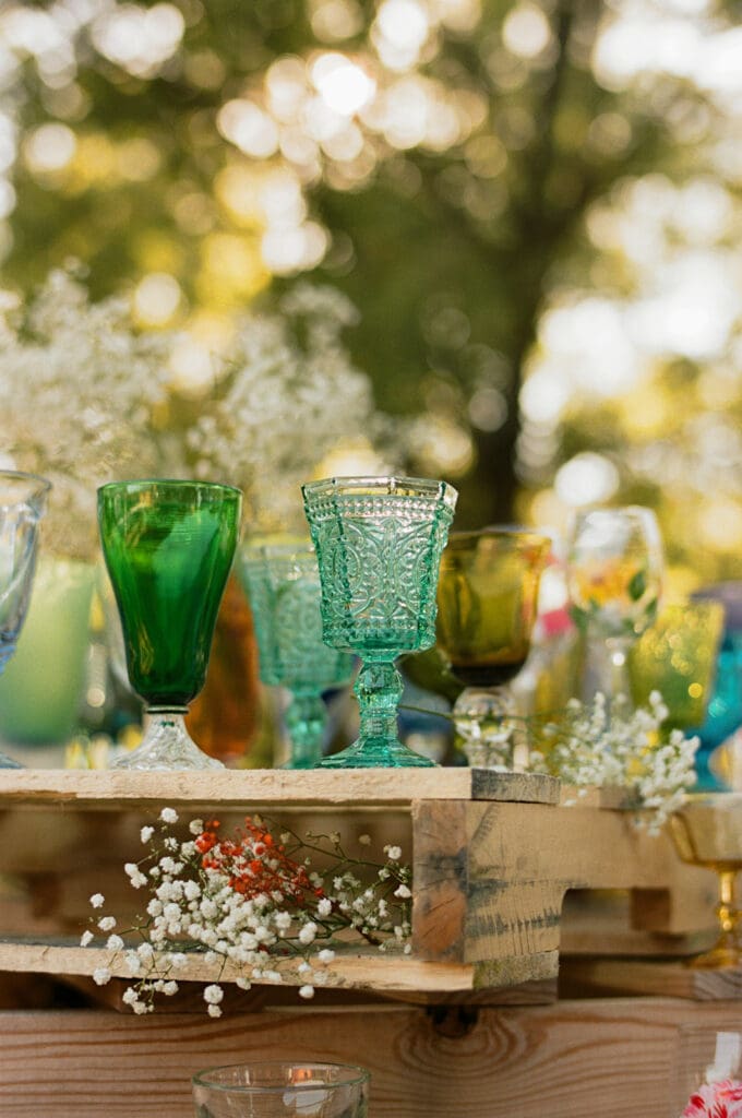 A variety of colorful glass goblets are arranged on a wooden pallet table, decorated with small white flowers, at an outdoor event.