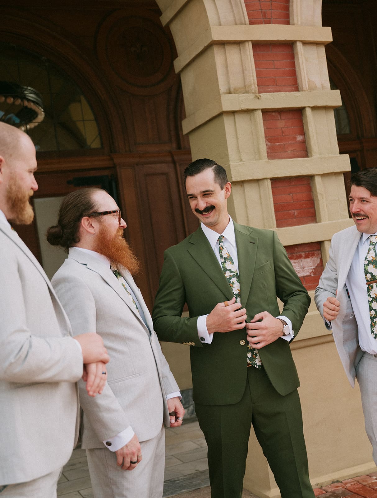 Four men in suits stand in front of an arched brick building entrance; three wear light gray suits and one wears a green suit.