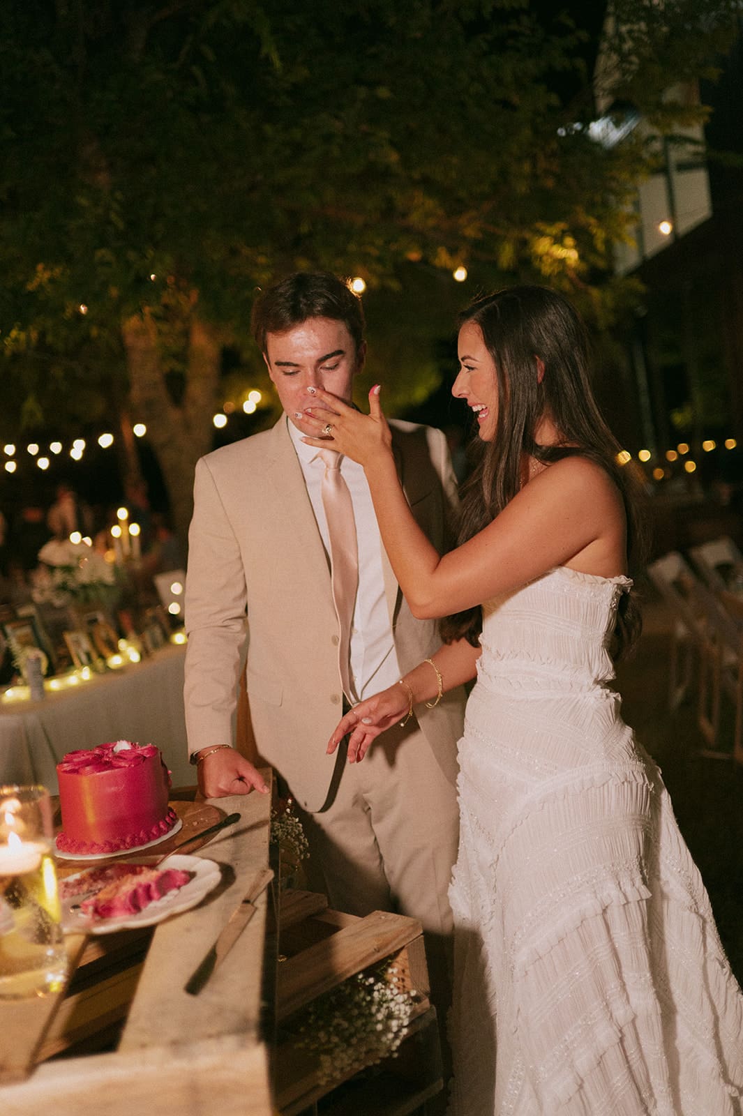 A group of people, including women in floral dresses, smile and dance outdoors at night under string lights for a backyard wedding 