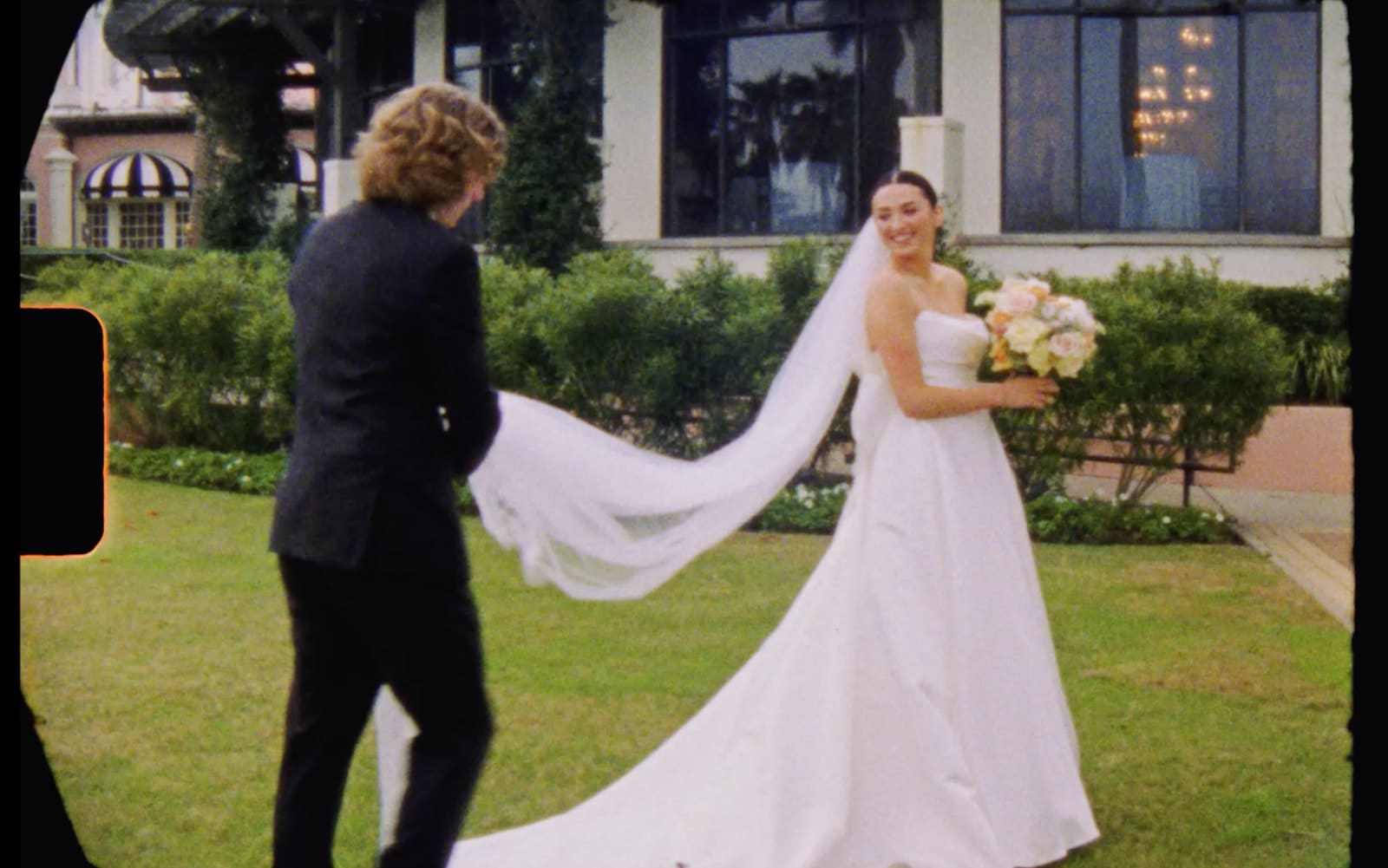 A bride and groom stand on a lawn in formal wedding attire, holding hands and smiling at each other, with a building and palm trees in the background on a supper 8mm wedding film