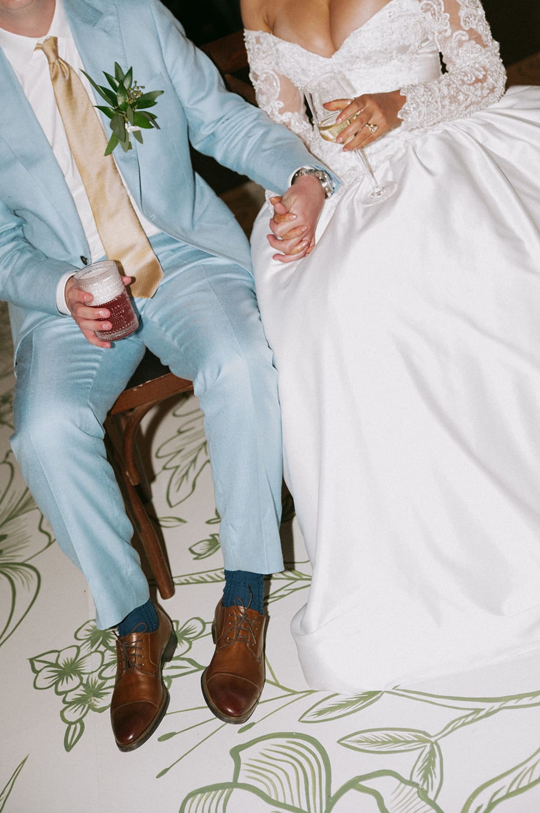 A bride in a white gown and a groom in a light blue suit sit side by side, holding hands. The groom holds a drink, and only their lower faces and outfits are visible.