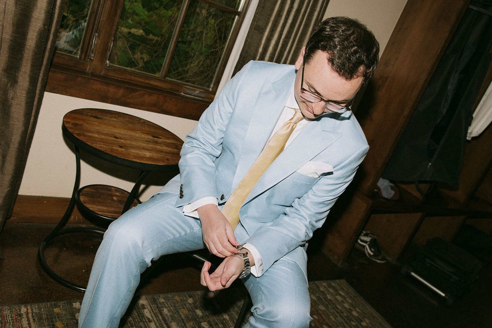 Man in a light blue suit and yellow tie sits on a bench indoors, adjusting his cufflink, with a window and round wooden table in the background.