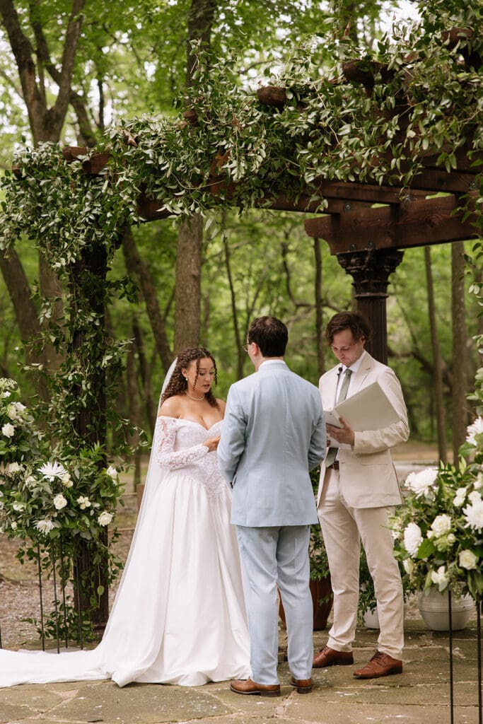 A bride and groom stand together at an outdoor wedding ceremony under a wooden pergola decorated with greenery and flowers, with two men officiating at The Springs Waxahachie