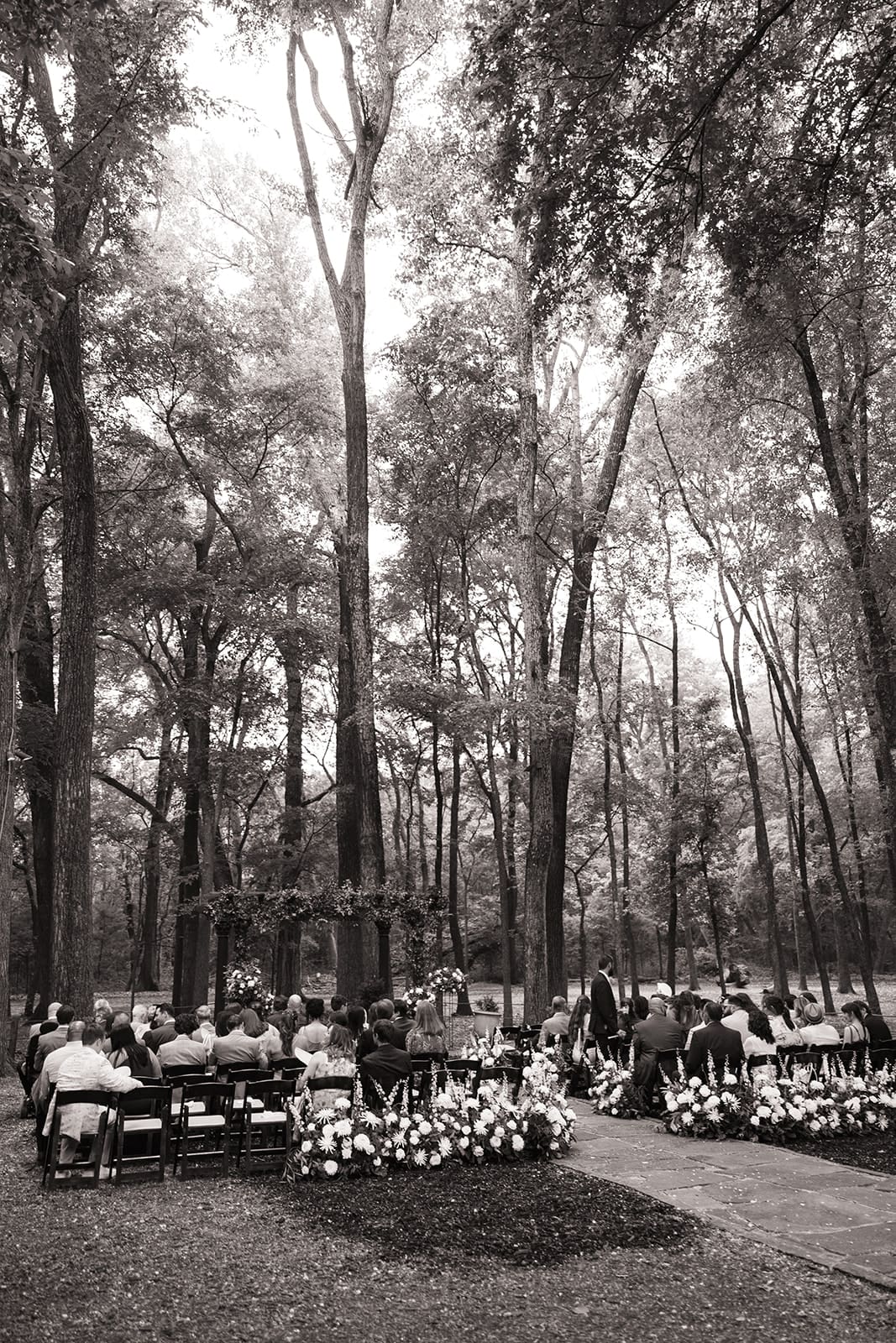 An outdoor wedding ceremony setup in a forest features a floral arch, potted plants, and white flowers lining the stone aisle, with trees in the background at The Springs Waxahachie