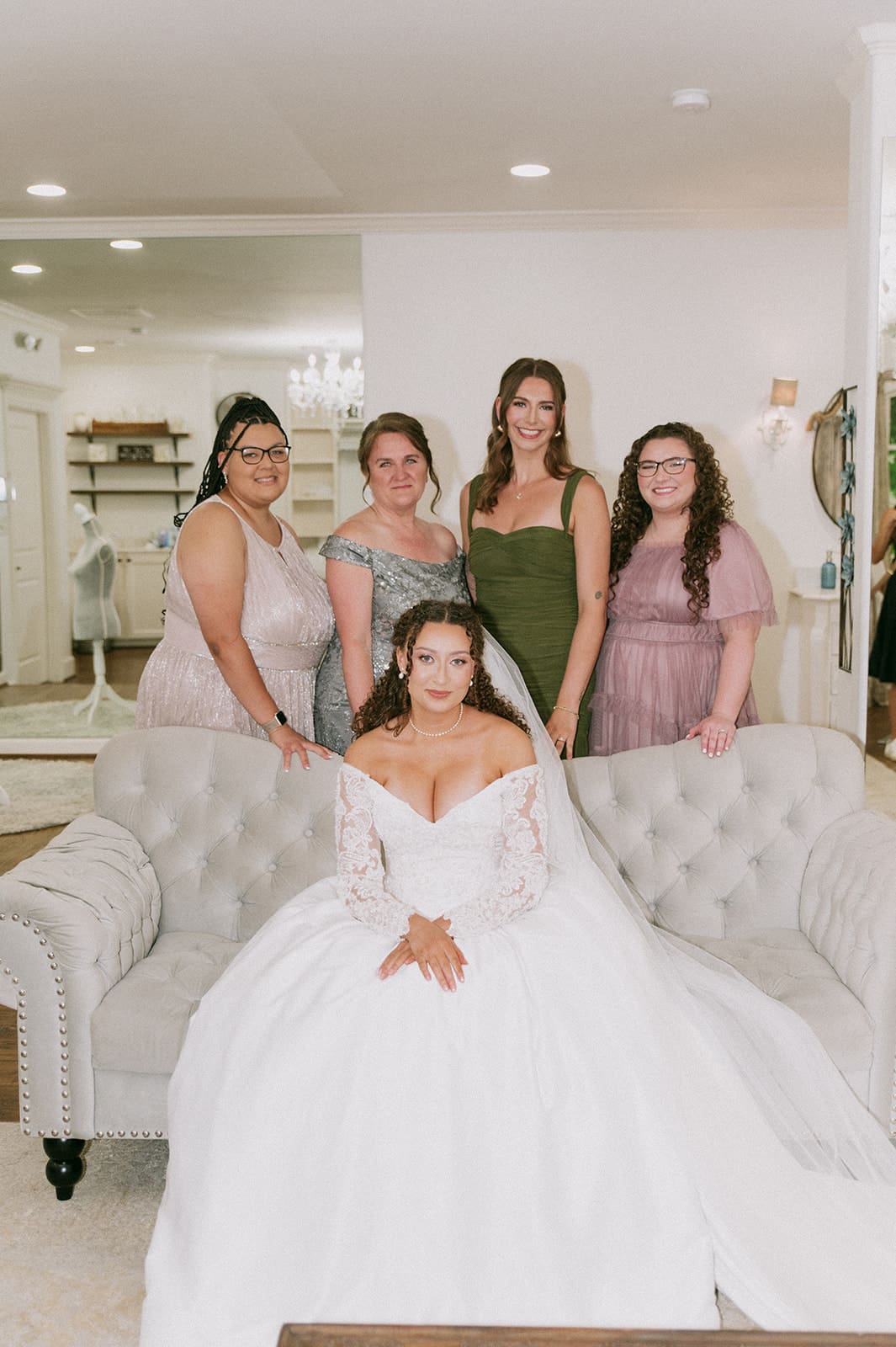 Five women pose in a bridal shop; one woman in a white wedding gown sits on a sofa while four others stand behind her, all wearing formal dresses for at The Springs Waxahachie