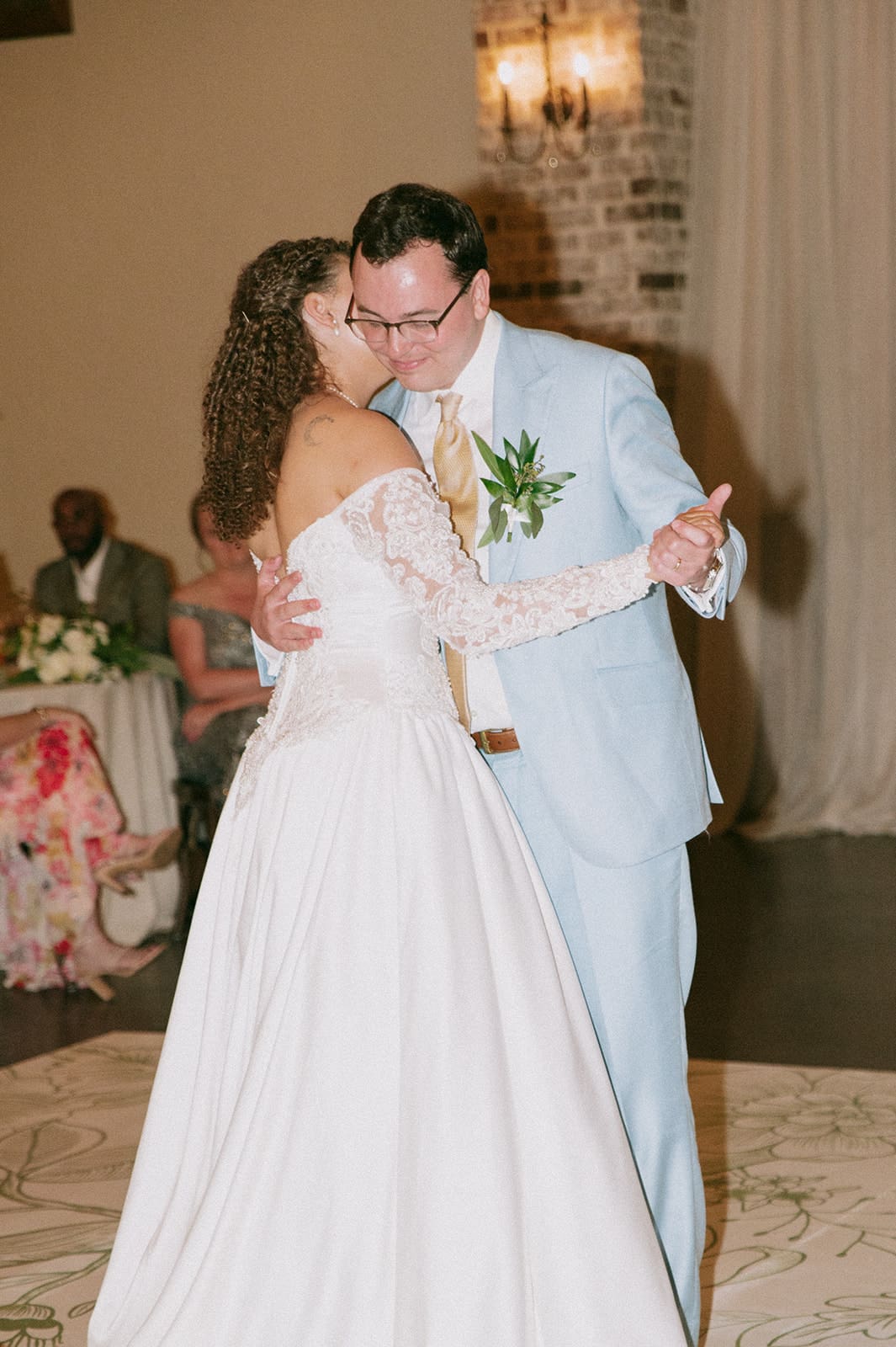 A bride in a white dress and a groom in a light blue suit share a dance at their wedding reception.