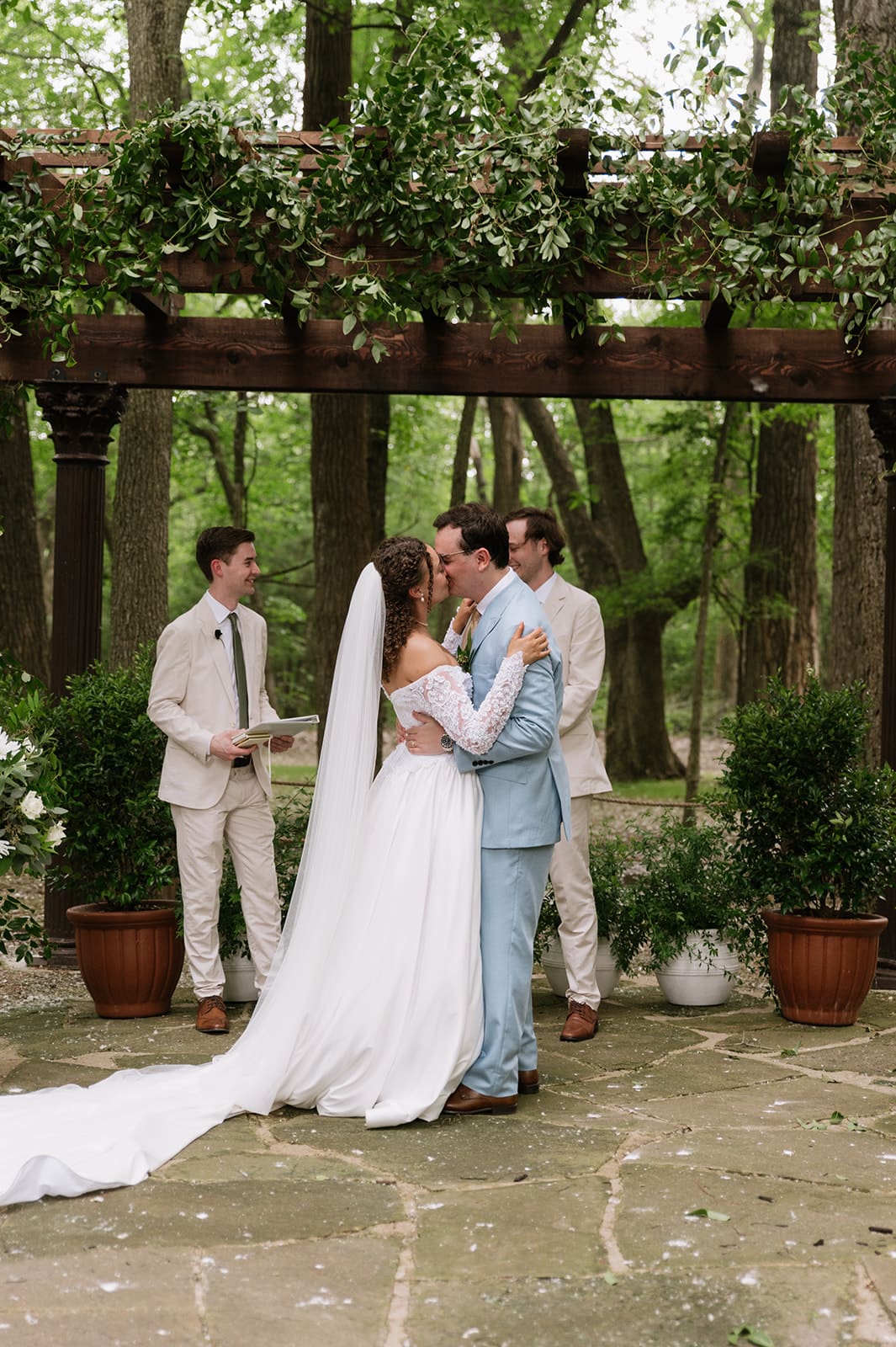 A bride and groom stand together at an outdoor wedding ceremony under a wooden pergola decorated with greenery and flowers, with two men officiating at The Springs Waxahachie