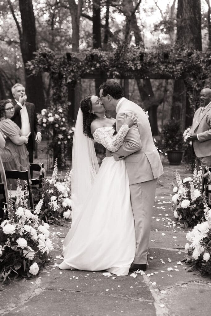 A bride and groom stand together at an outdoor wedding ceremony under a wooden pergola decorated with greenery and flowers, with two men officiating at The Springs Waxahachie