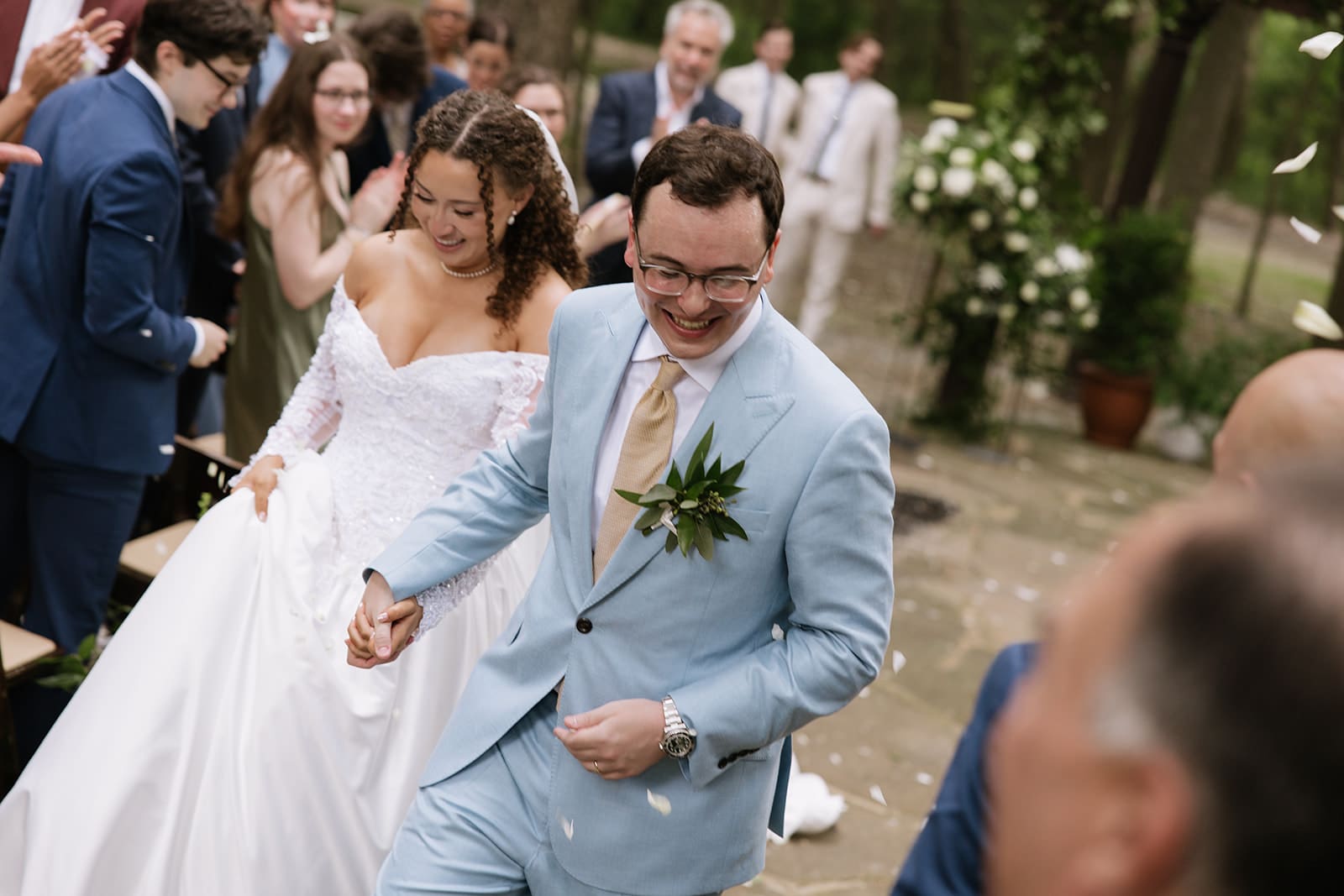 A bride in a white dress and a groom in a light blue suit walk hand in hand outdoors among guests, smiling as petals are thrown.