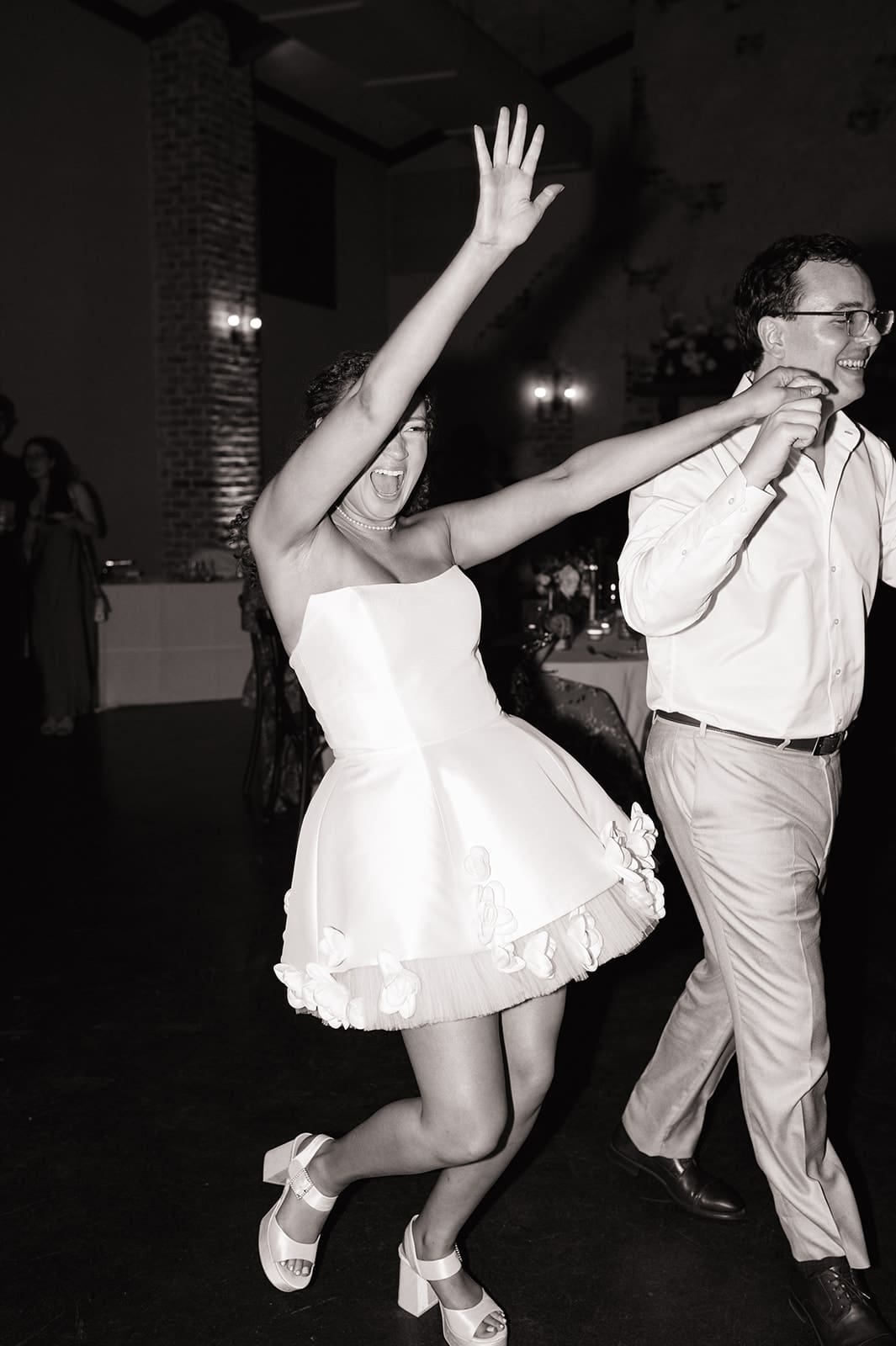 A woman in a short white dress and a man in light blue pants and a white shirt hold hands and dance indoors at a formal event.