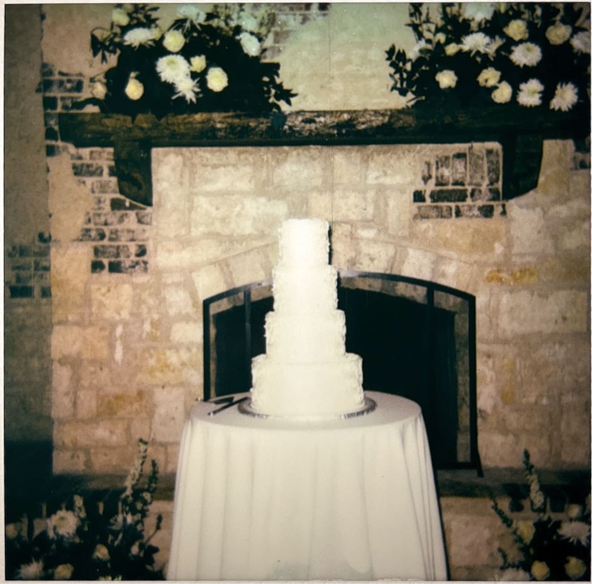A three-tiered white wedding cake sits on a round table in front of a stone fireplace decorated with white flowers.