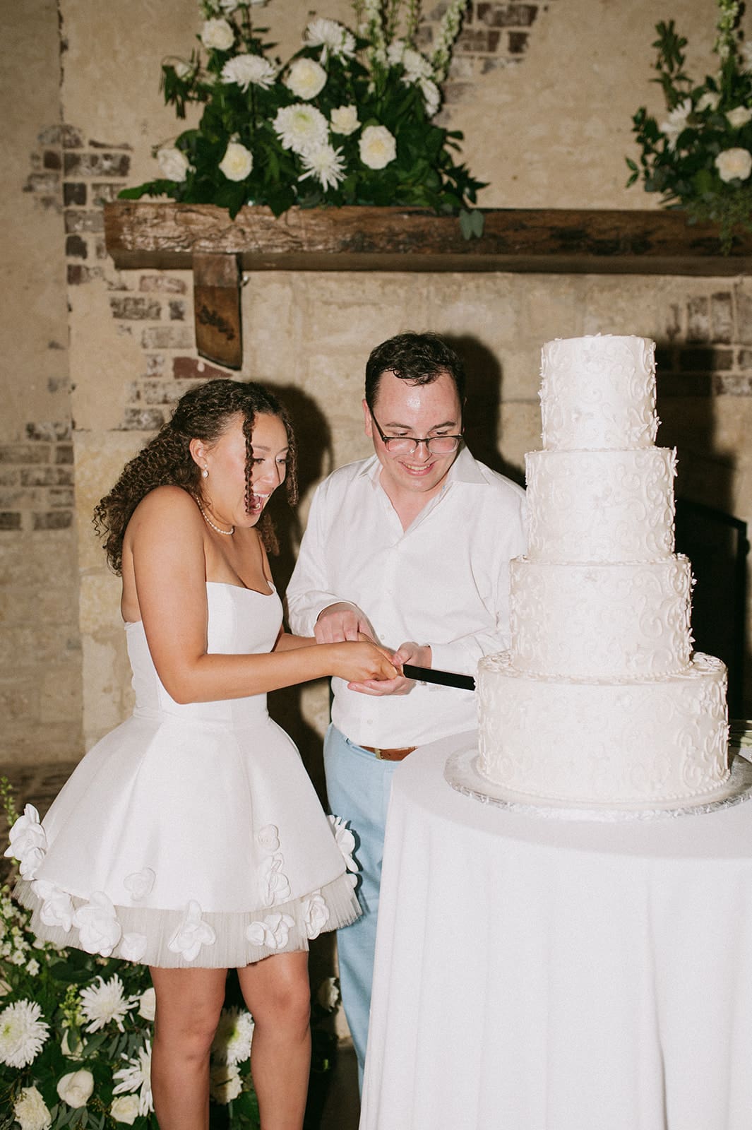 A three-tiered white wedding cake sits on a round table in front of a stone fireplace decorated with white flowers.