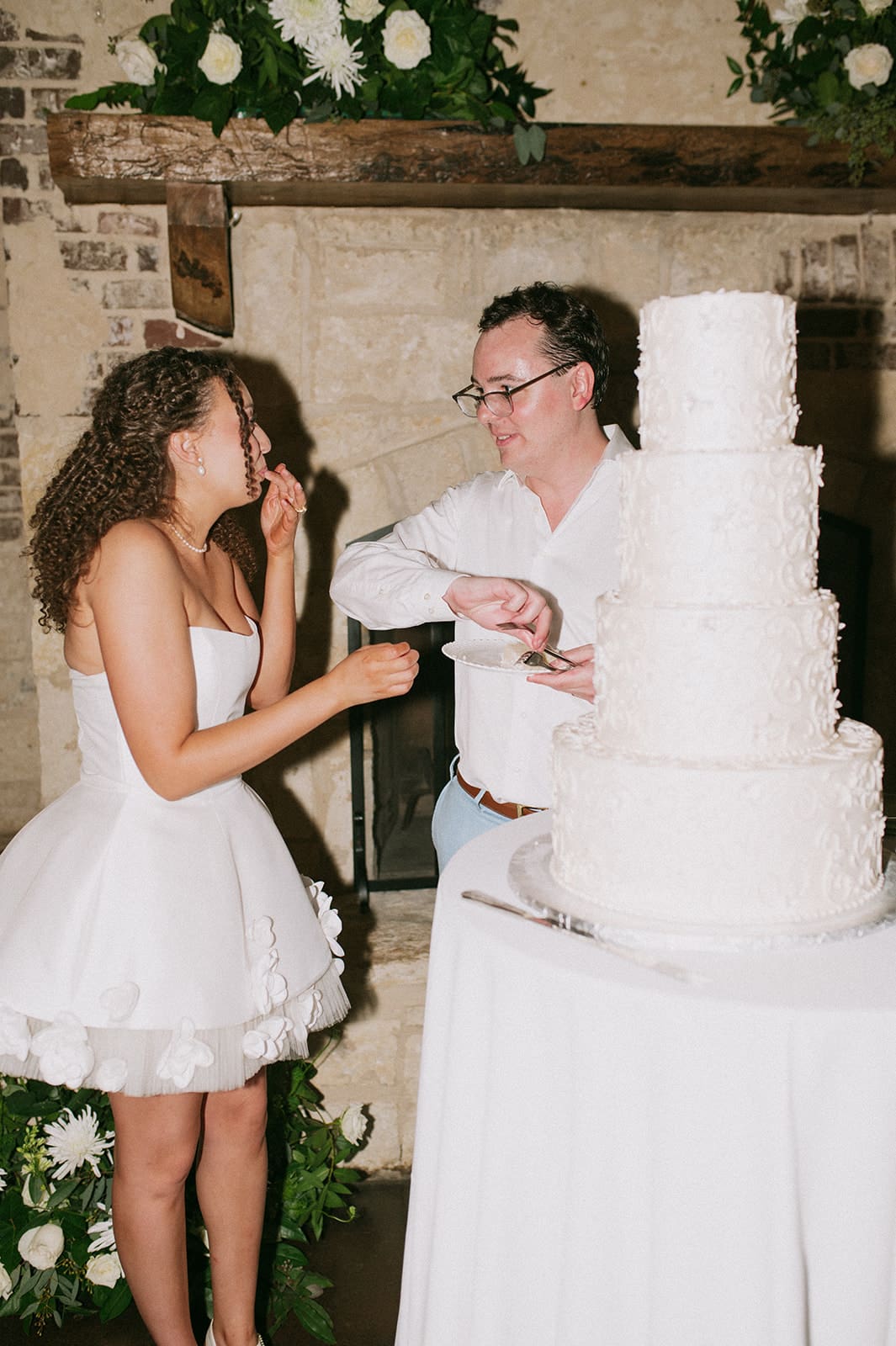  A three-tiered white wedding cake sits on a round table in front of a stone fireplace decorated with white flowers.