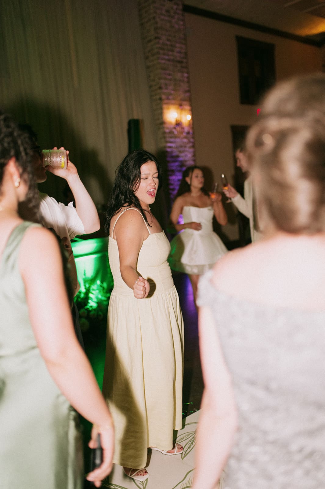 A group of people in formal attire dance together at an indoor event. One woman in a light dress stands in the center, reaching out her hand.