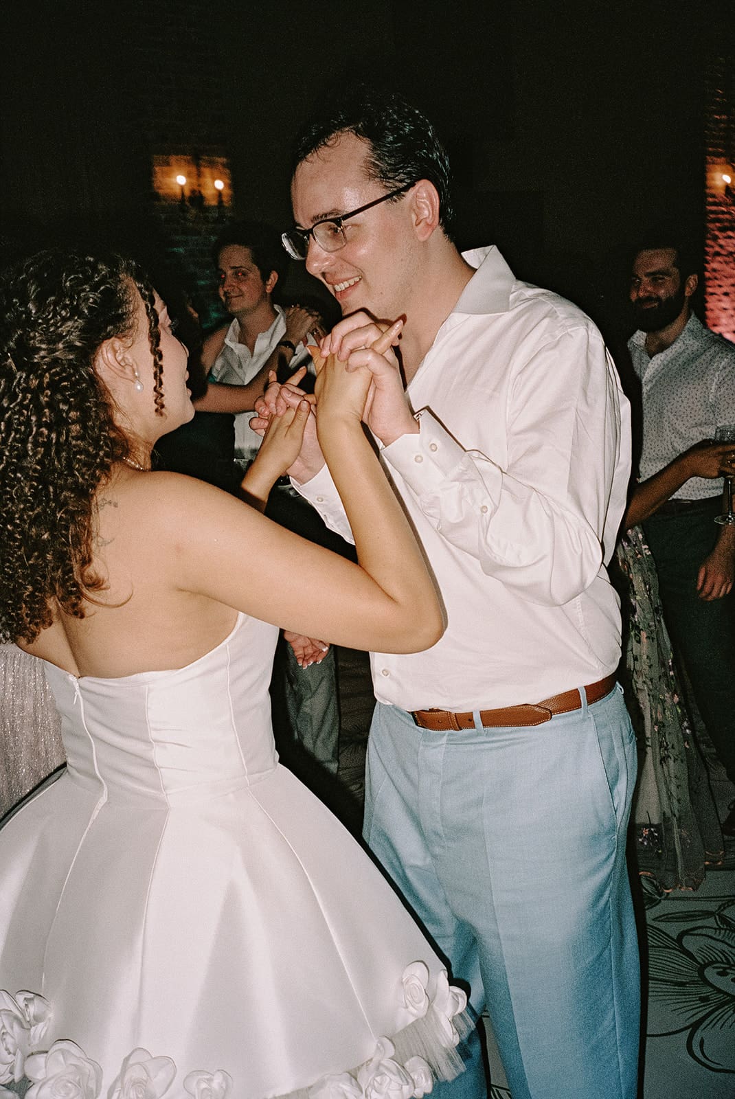 A group of people in formal attire dance together at an indoor event. One woman in a light dress stands in the center, reaching out her hand.